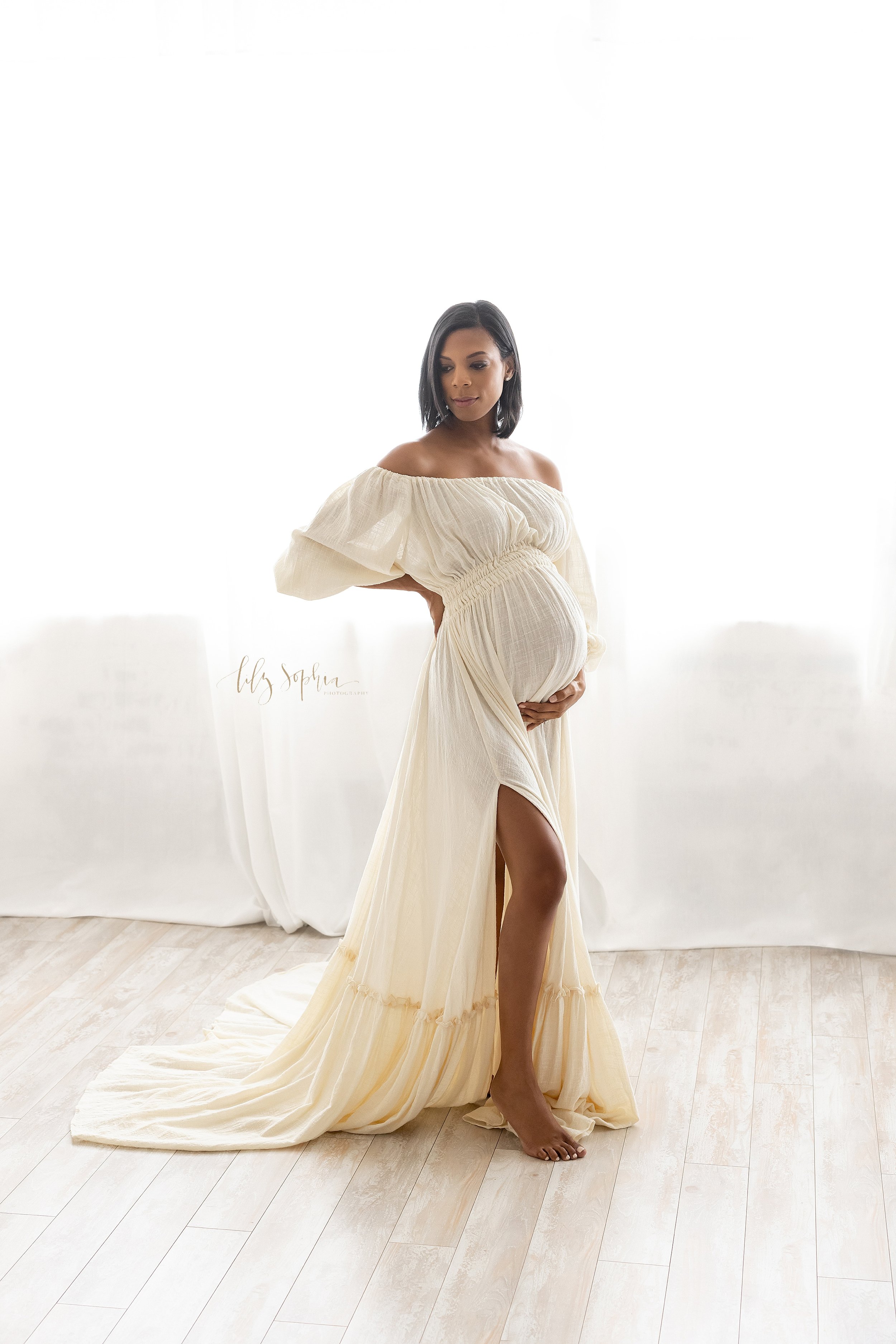  Maternity portrait of an African-American pregnant mother wearing a peasant off-the shoulder full-length gown as she looks over her right shoulder while placing her right hand on her hip and her left hand at the base of her belly  as she stands in f