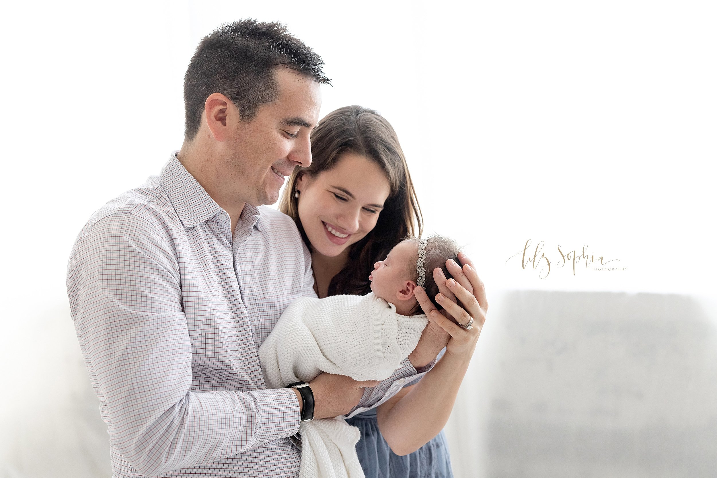  Newborn photo shoot in front of a window streaming natural light of a father holding his newborn baby girl in his hands in front of his chest while mom stands on his left side and the two of them admire their daughter taken in a studio near Ansley P