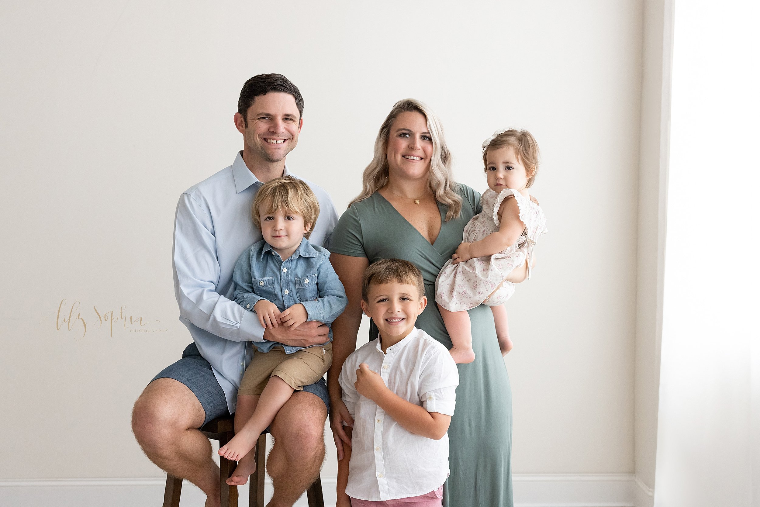  Family portrait of a father sitting on a stool and holding his toddler son as his wife stands to his left side holding their one year old daughter and their older son stands in front of mom taken in natural light in a studio near Sandy Springs in At