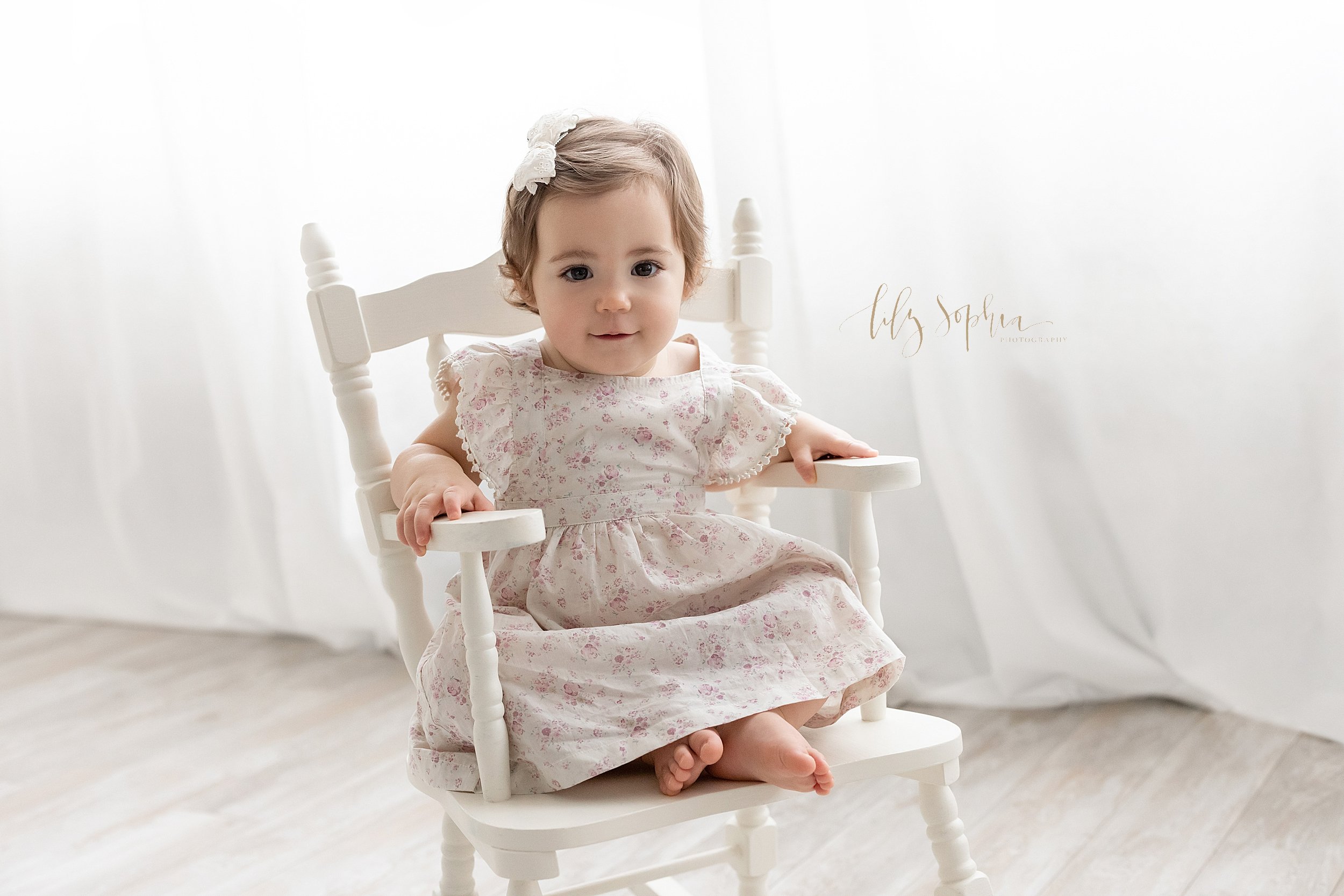  First birthday portrait of a one year old girl as she sits in a wooden rocking chair in front of a window streaming natural light in a photography studio near  Alpharetta in Atlanta. 