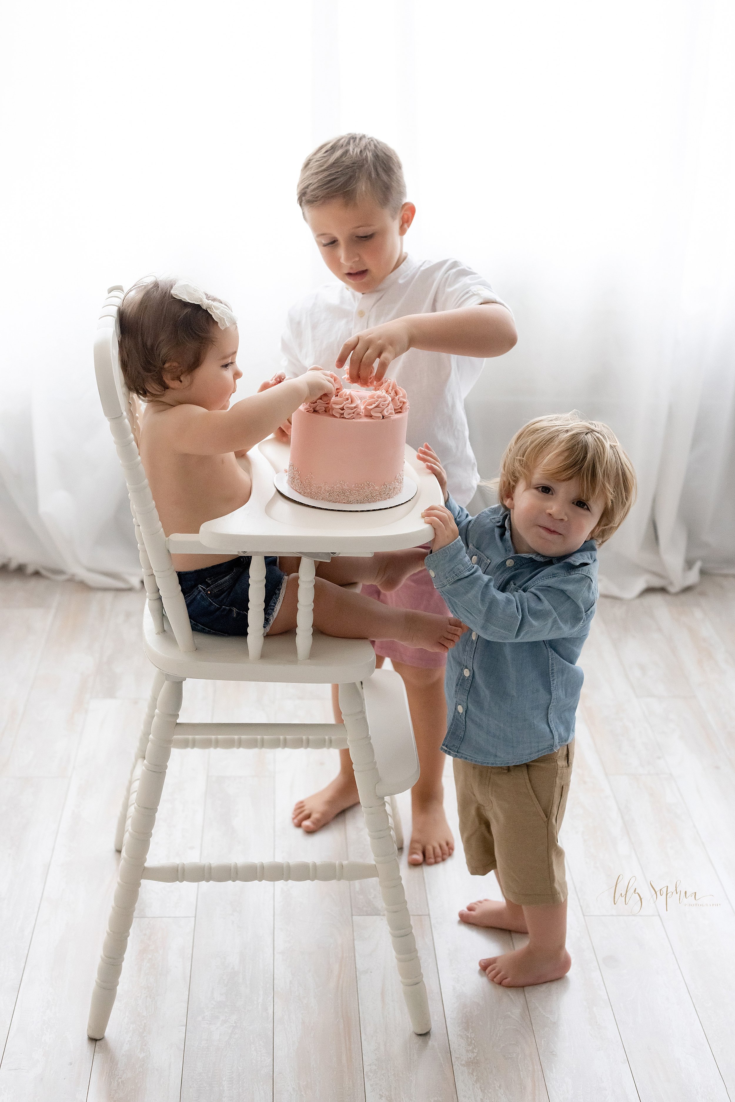  First birthday smash cake photo of a one year old baby girl sitting in an antique high chair grabbing icing on her cake as her older brothers try to help taken in front of a window streaming natural light in a studio near Virginia Highlands in Atlan