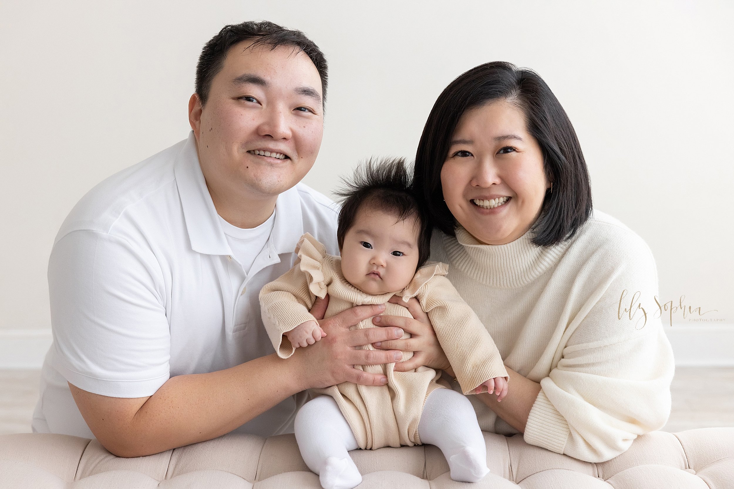  Family portrait of an Asian mother and father as they kneel behind a tufted upholstered bench and support their baby girl as she sits atop the bench taken in a natural light studio near Midtown in Atlanta, Georgia. 