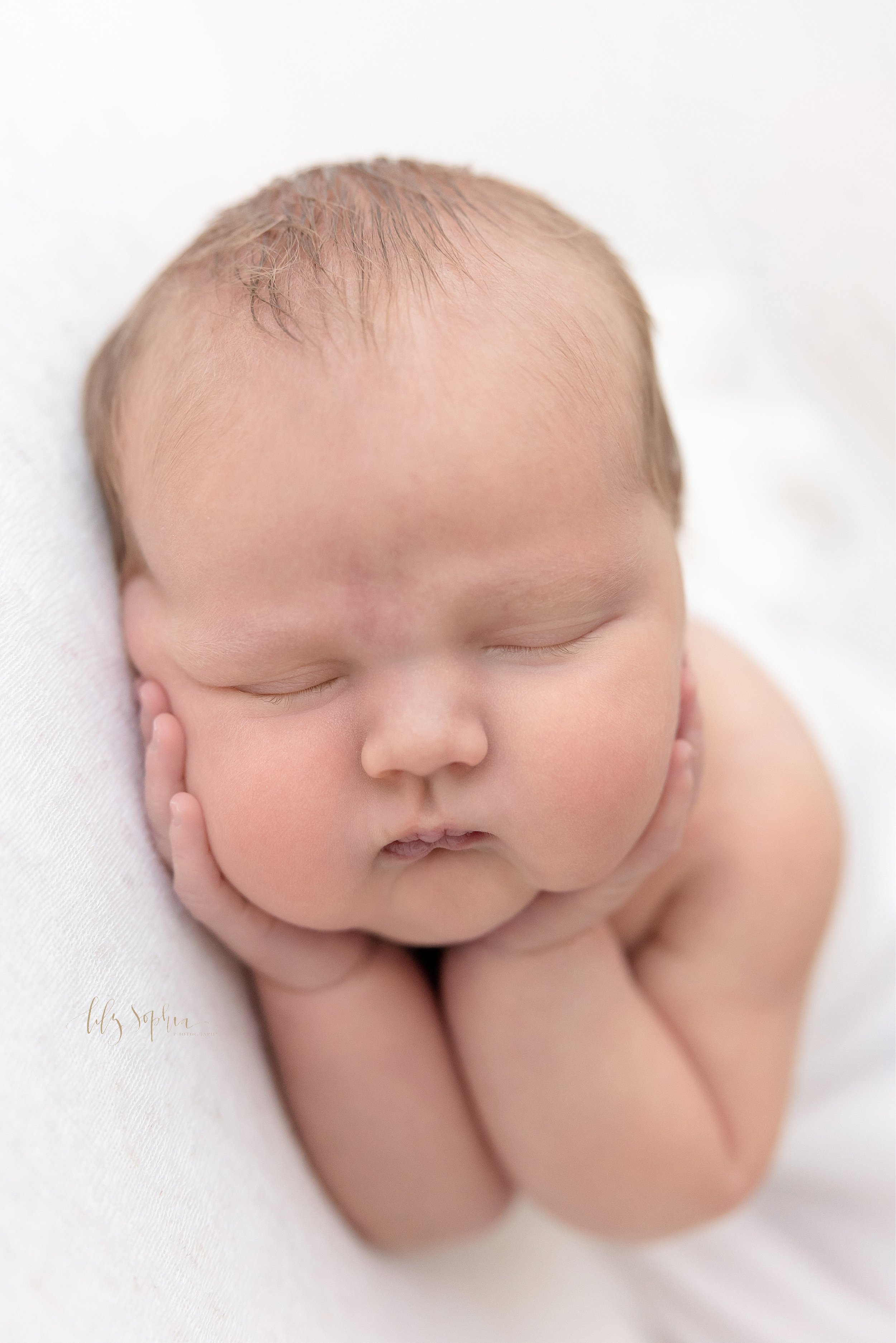  Newborn photo shoot of a newborn baby boy peacefully sleeping as he holds his cheeks in his hands taken in a studio near Cumming in Atlanta that uses natural light. 