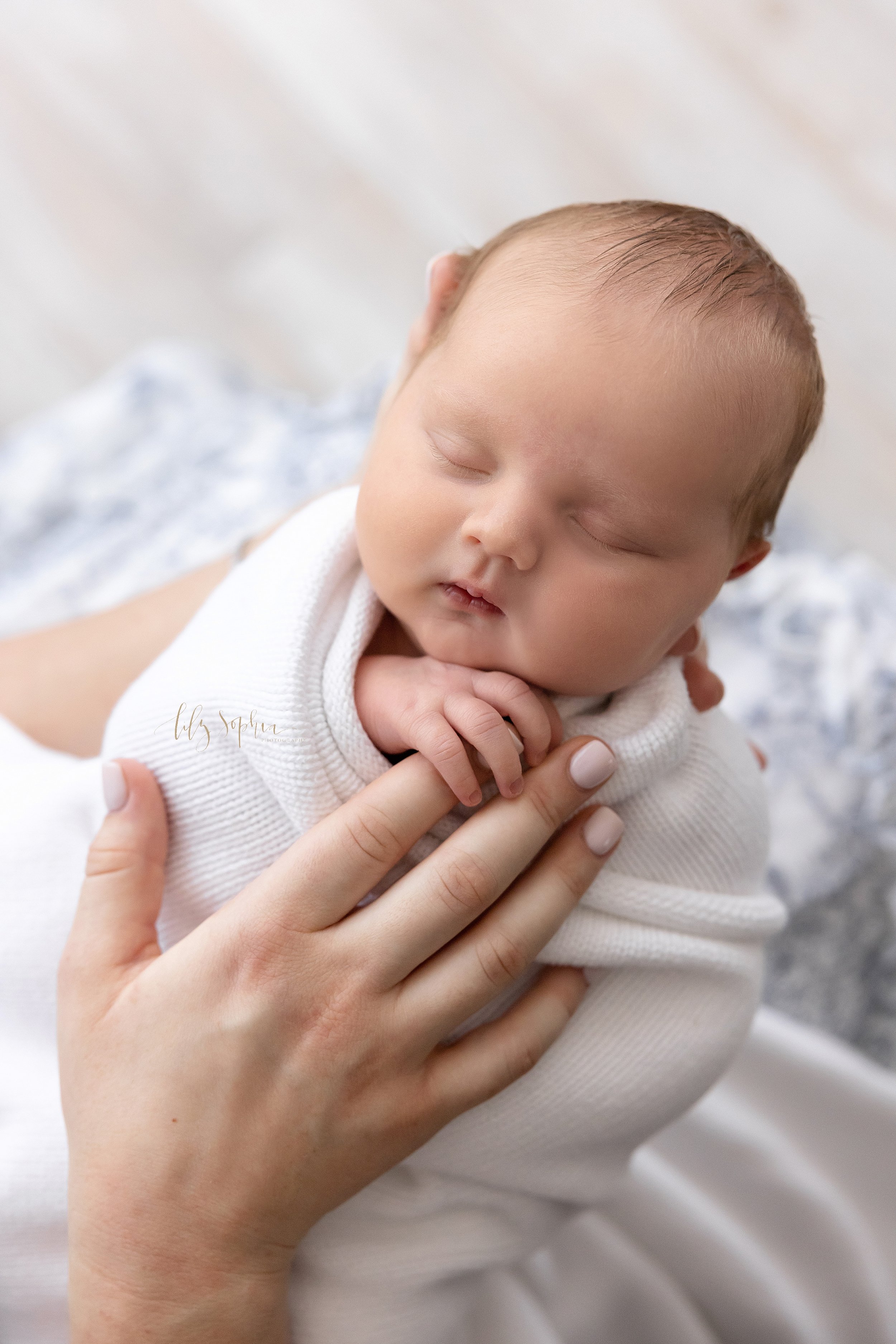  Newborn portrait of an infant boy swaddle to his chin in a white blanket as he sleeps with his head in his mother’s hand and the baby boy holds onto his mother’s index finger taken near Kirkwood in Atlanta in a studio using natural light. 