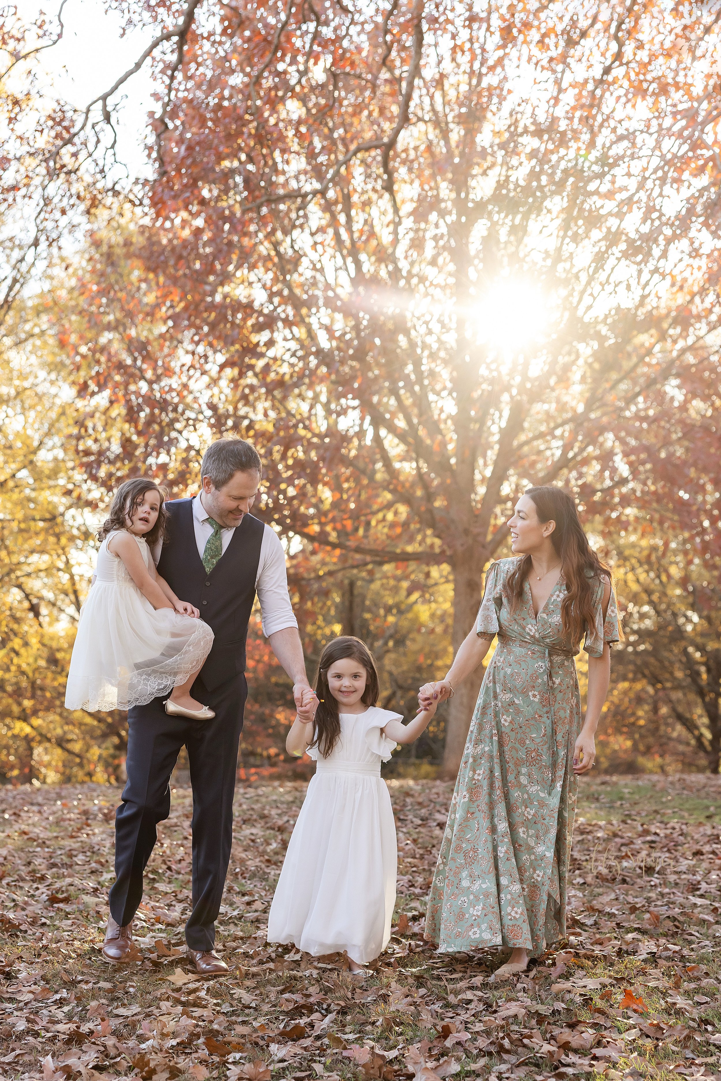  Family photo shoot in an Atlanta park of a father holding his youngest daughter in his right arm as he he and his wife walk holding hands with their older daughter between them during autumn at sunset; 