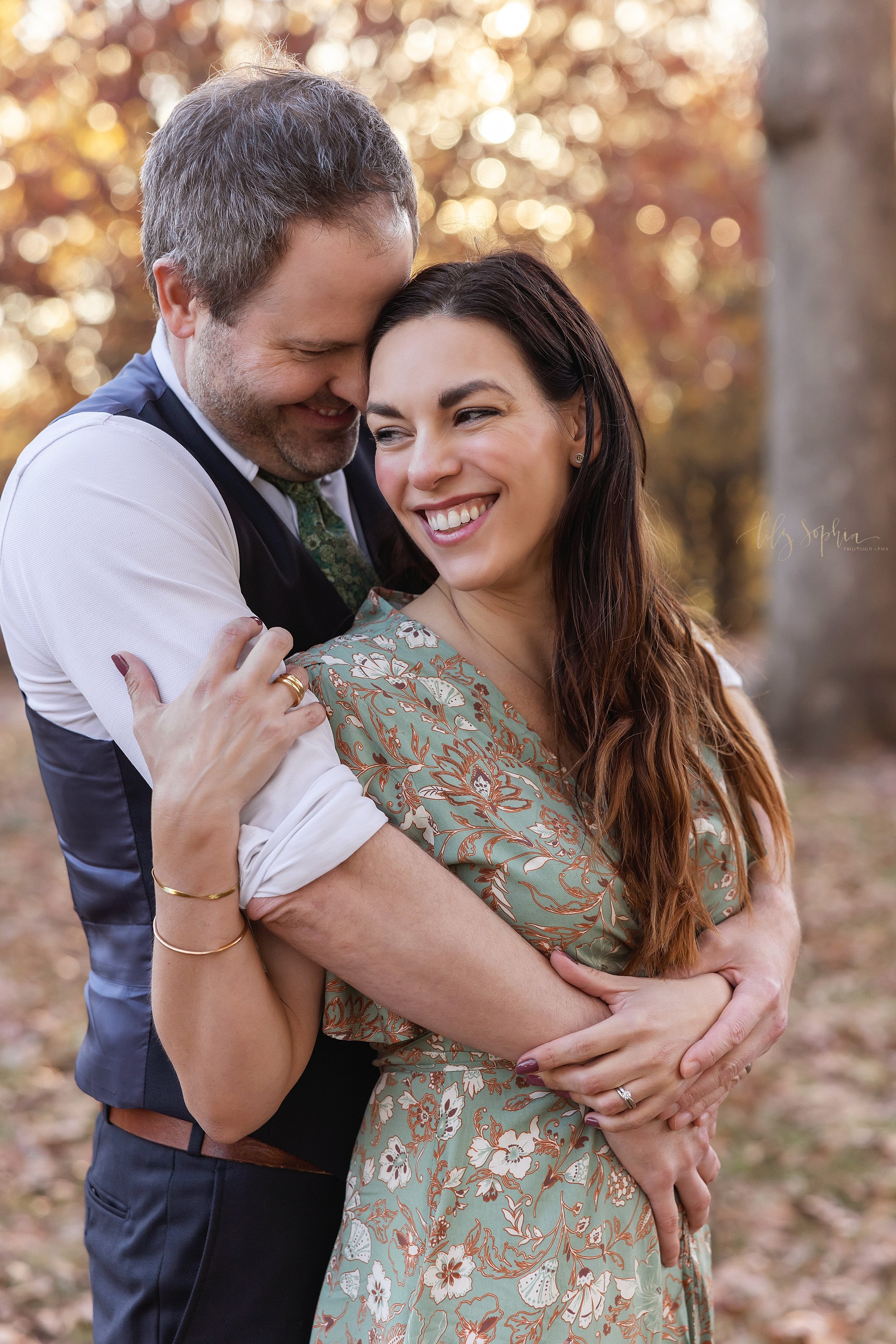  Family picture of a husband wrapping his arms around his wife as she stands in front of him and turns her head to look over her right shoulder as the couple stand among the autumn leaves at sunset in an Atlanta park. 