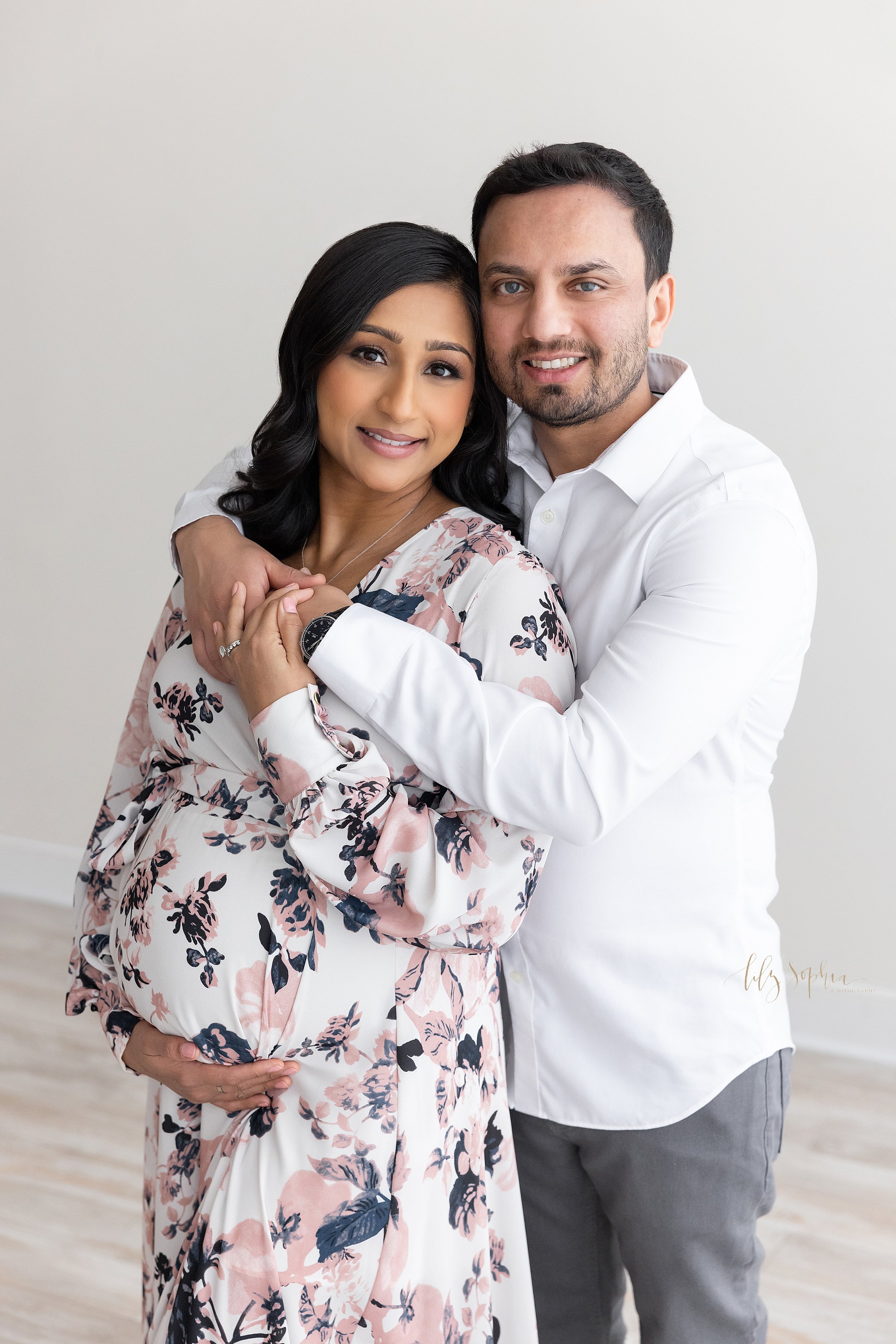  Maternity photo shoot with a pregnant Indian mother standing with her right hand holding the base of her belly and her back against her husband’s chest as he wraps his arms around her taken in a photography studio that uses natural light near Brookh