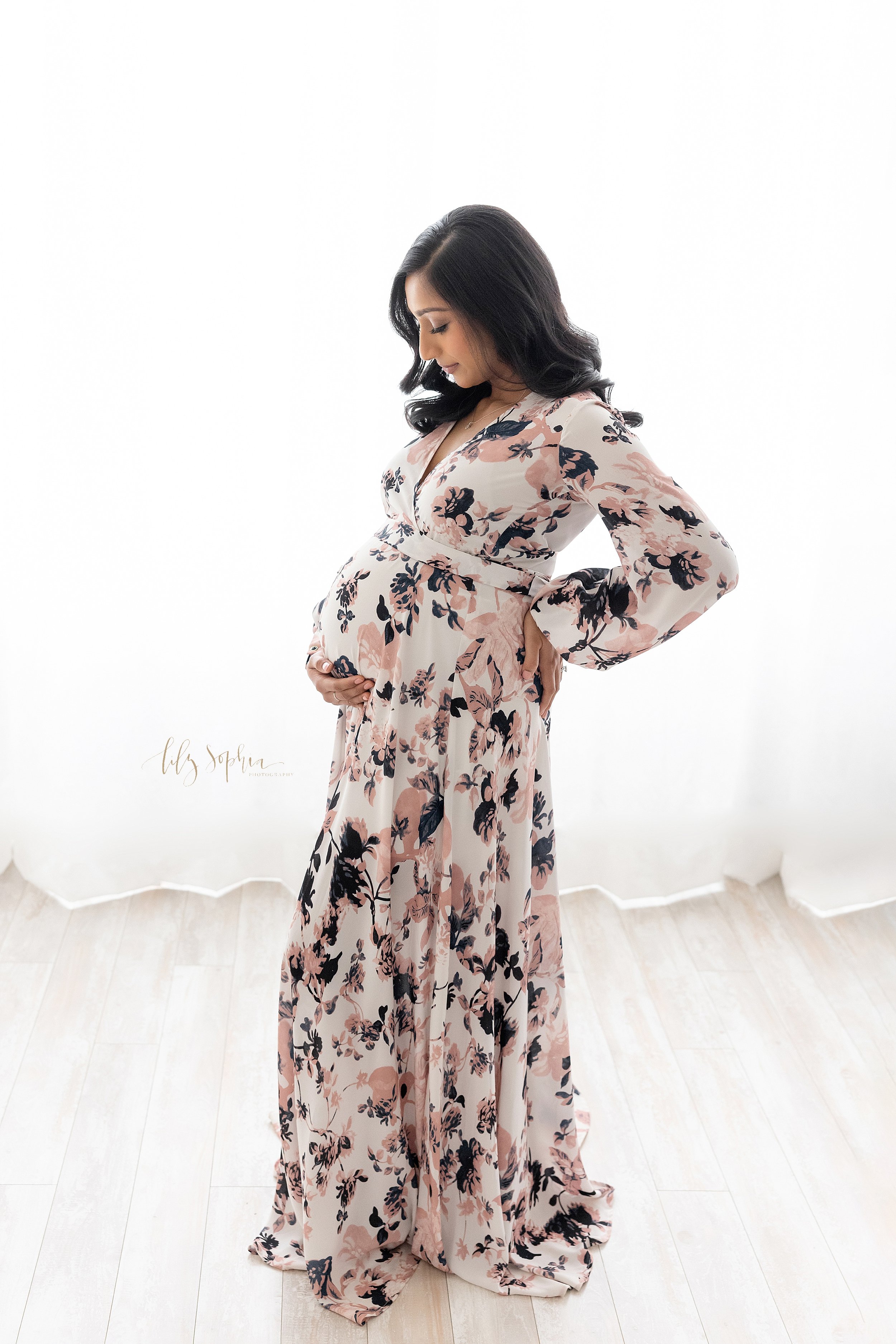  Maternity portrait of an Indian pregnant mother as she stands in front of a window streaming natural light wearing a floral long sleeve full-length gown with her right hand holding her belly and her left hand on her hip as she contemplates the upcom