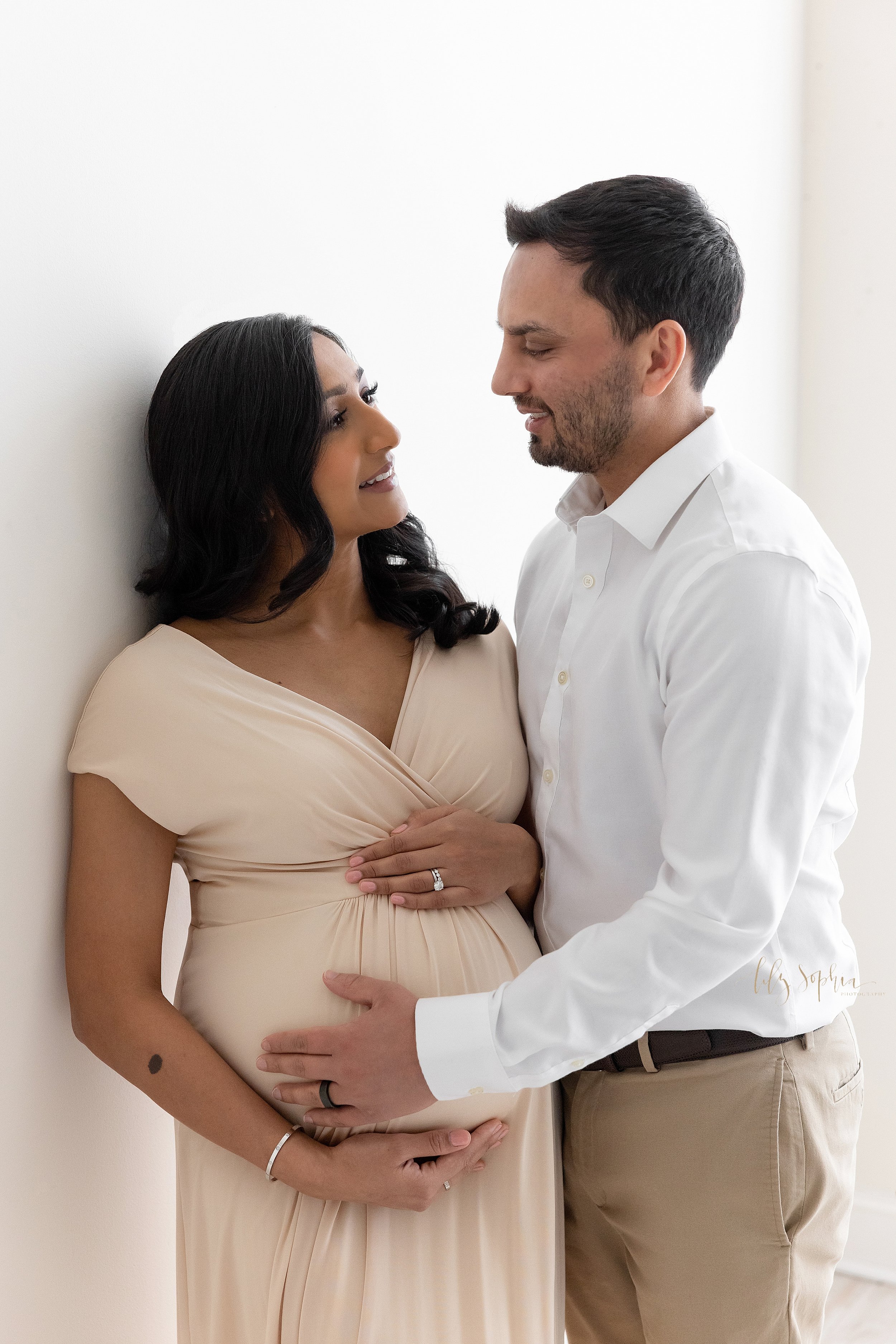  Maternity photo of an Indian expectant couple with the wife leaning against a wall with her right shoulder as she frames her belly with her hands and her husband faces her on her left side and places his right hand on their child in utero while they