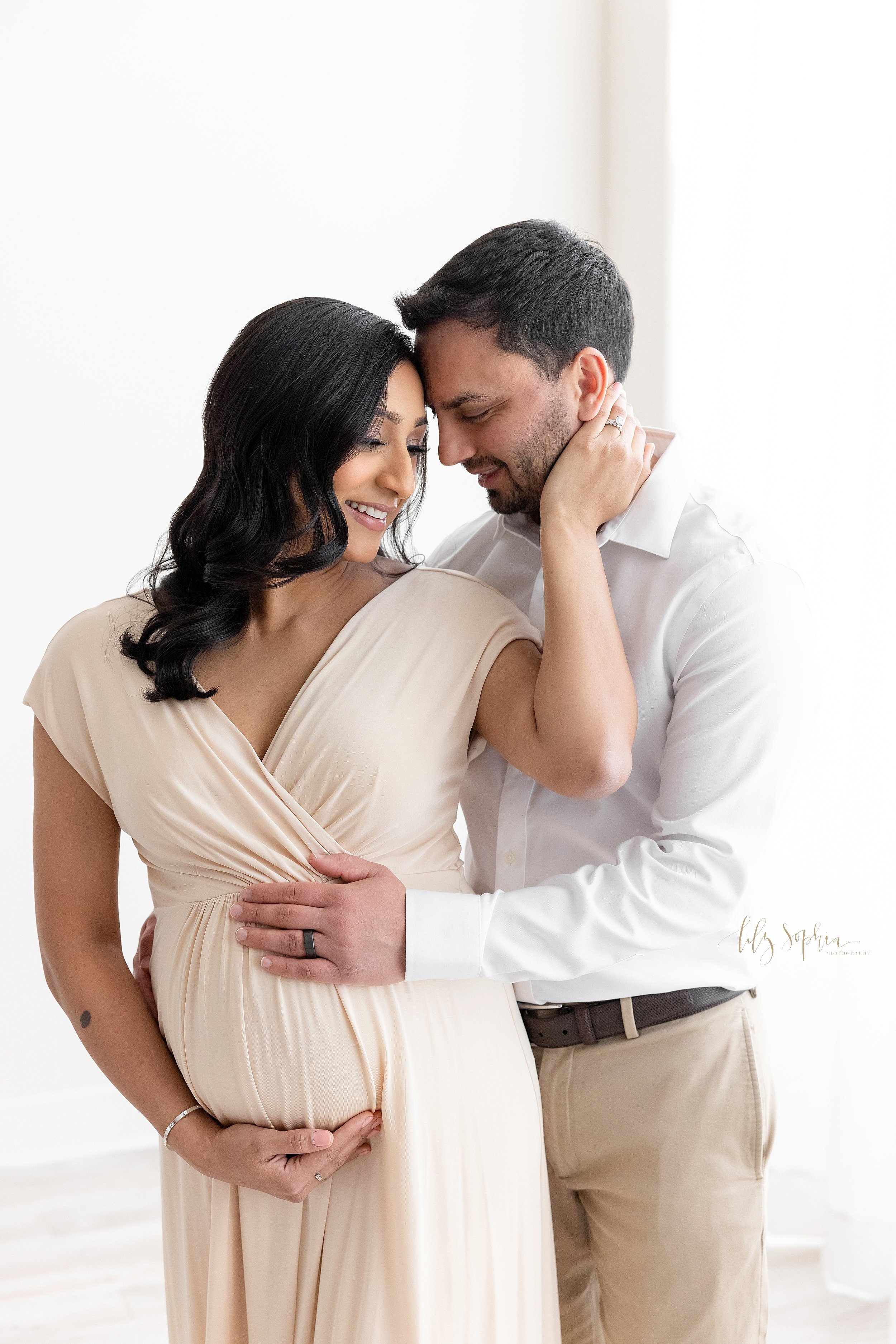 Maternity photo shoot with a pregnant Indian mother as she stands with her right hand holding the base of her belly and her left hand holding the face of her husband while he stands to her left and wraps his arms around his wife’s waist taken in fro