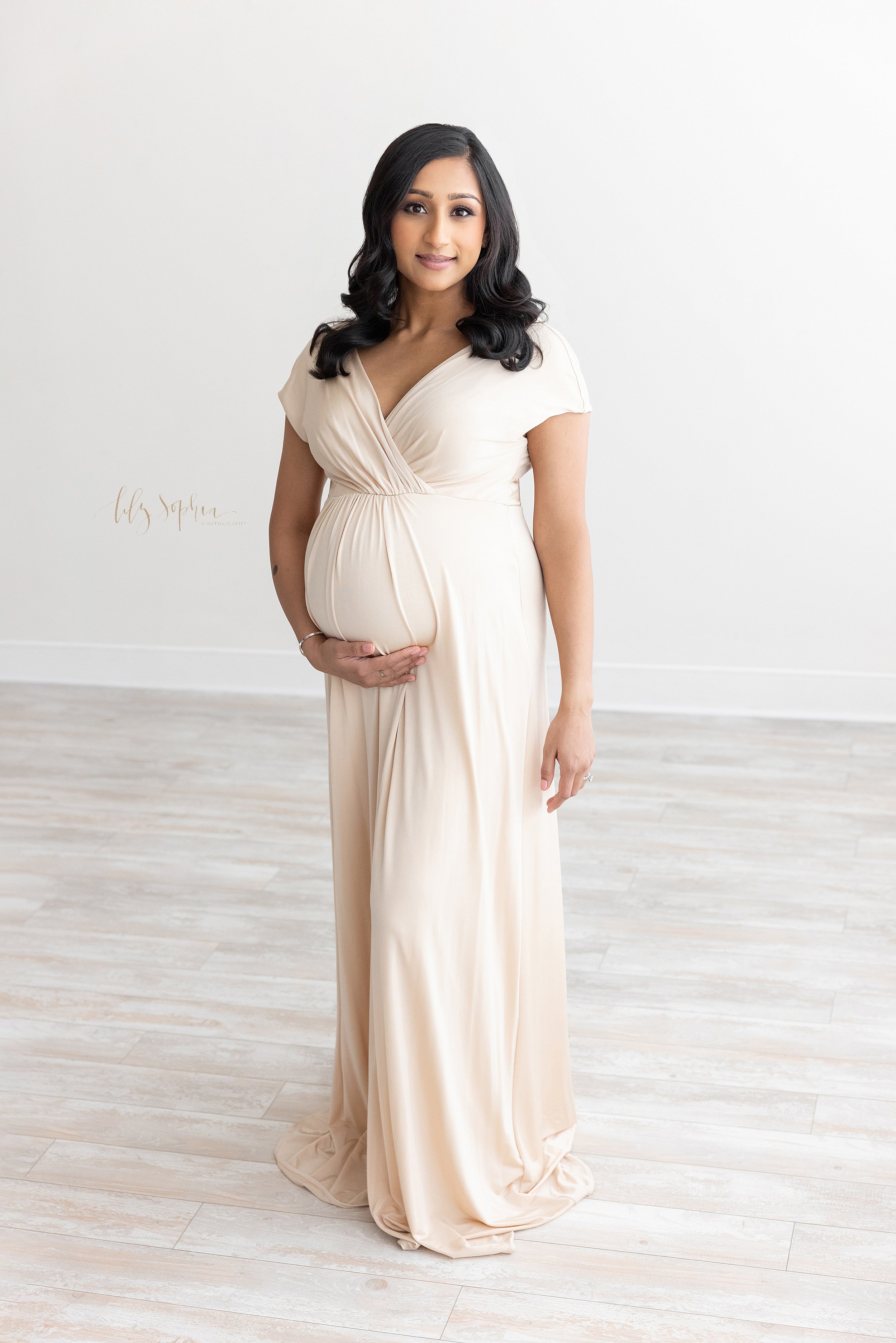  Maternity portrait of a pregnant Indian mother as she stands holding her belly with her right hand in a studio that uses natural light near Old Fourth Ward in Atlanta. 