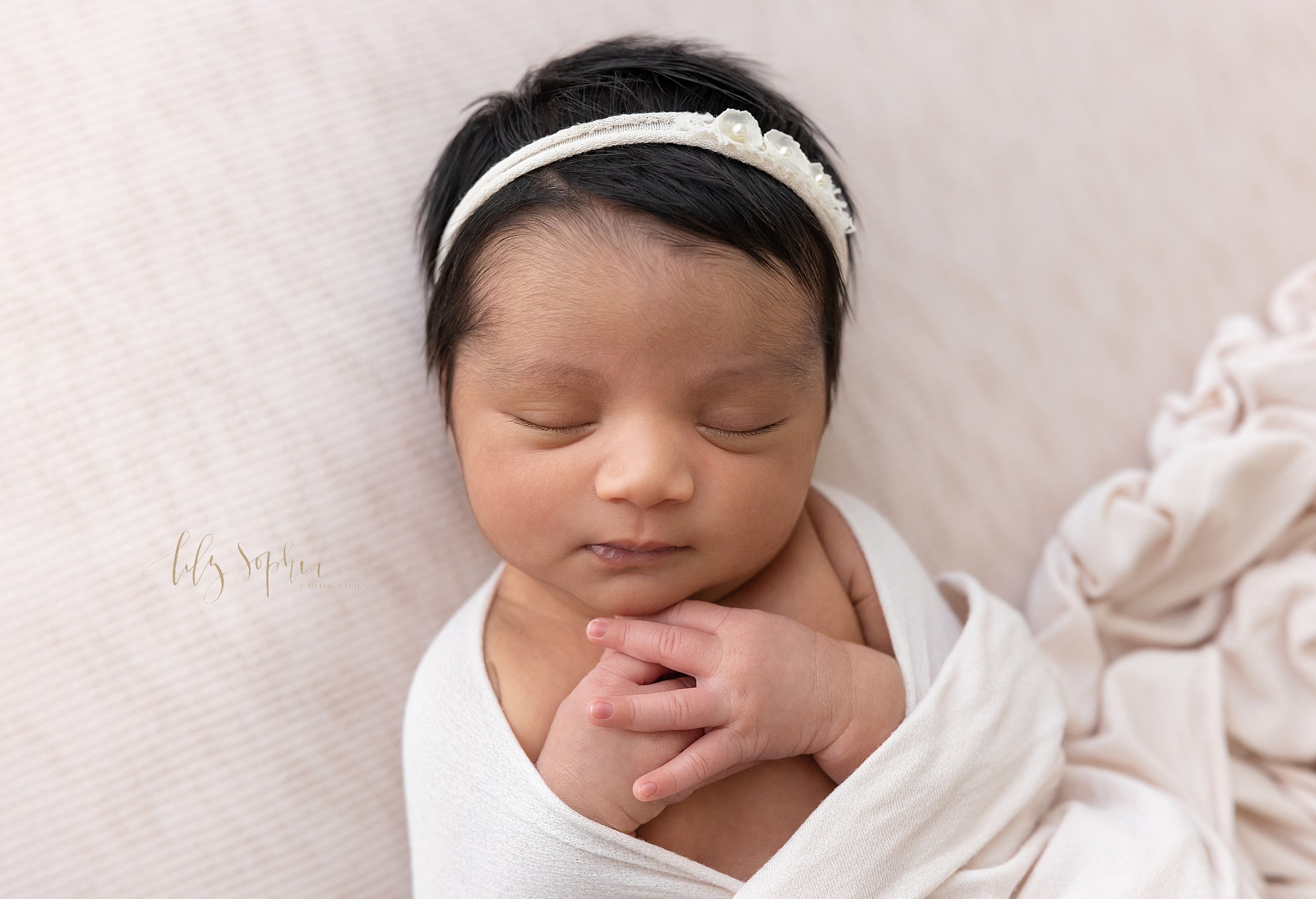  Newborn portrait of a sleeping Indian newborn baby girl as she is draped in a soft stretchy blanket, wearing a delicate headband in her jet black hair and holding her hands together on her chest taken near Midtown in Atlanta in natural light at a ph