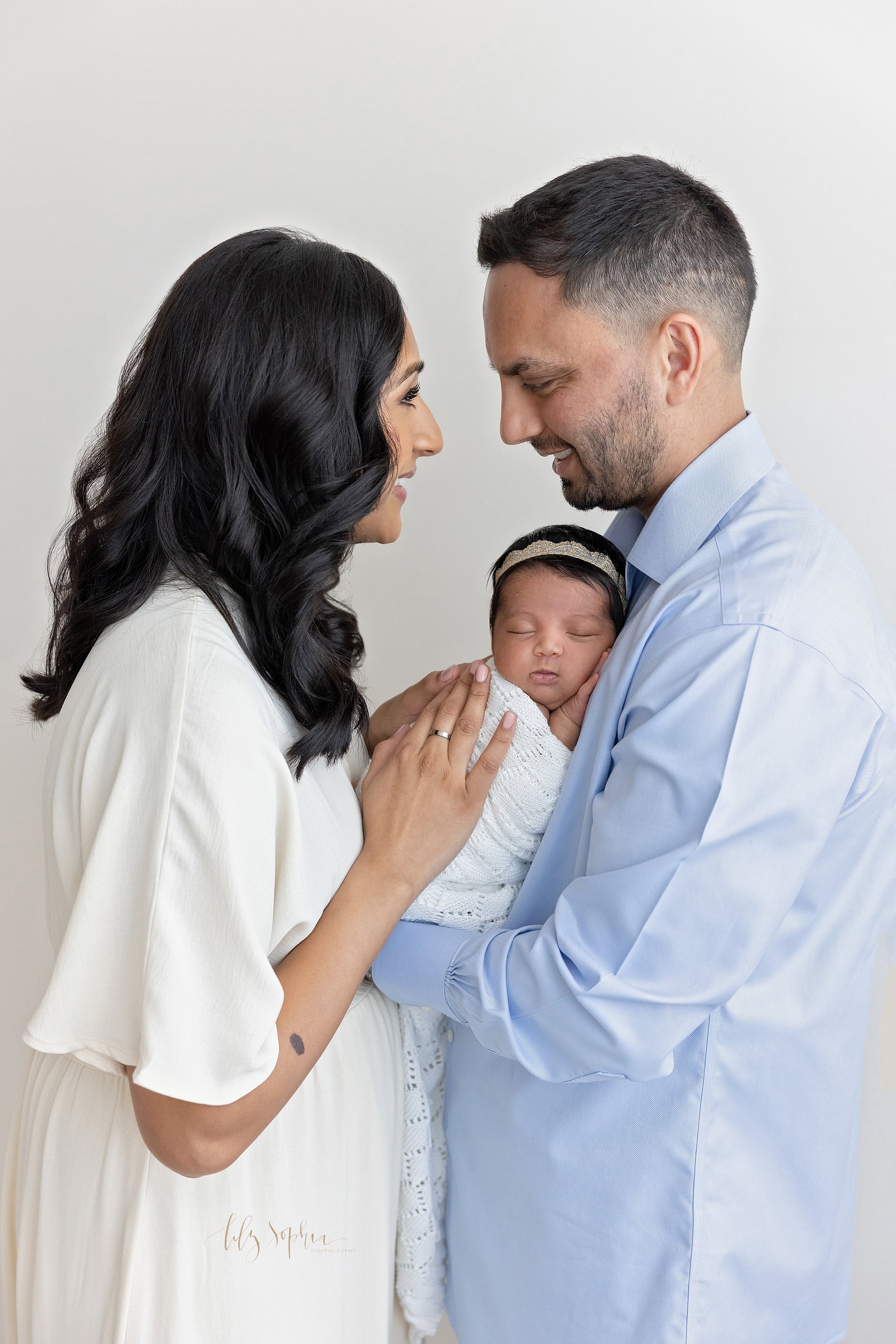  Newborn family photo of an Indian father holding his sleeping newborn baby daughter against his chest as his wife faces him and places her hands on their daughter’s back while the parents smile lovingly at one another taken in natural light in a pho