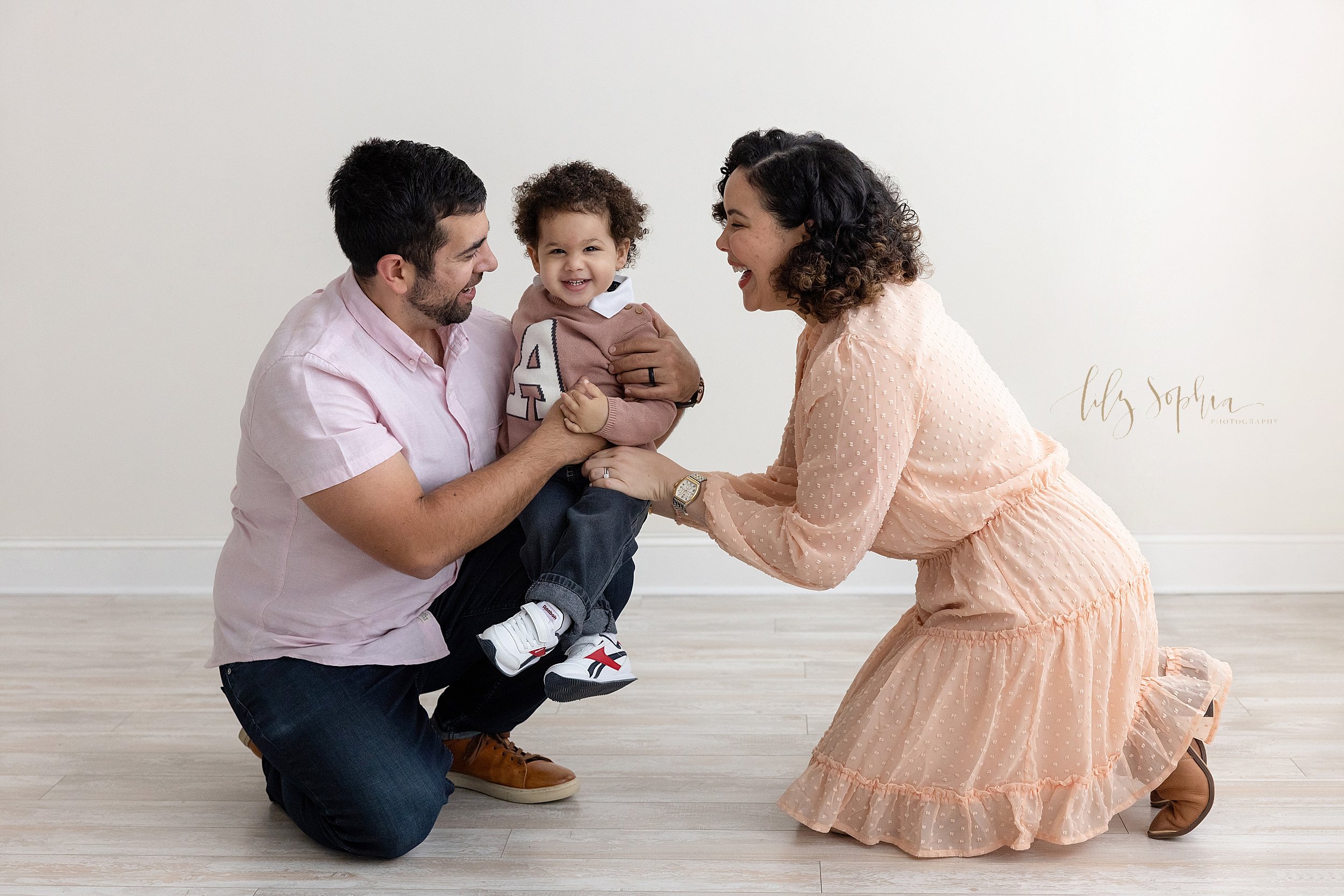  Family photo shoot in a natural light studio near Oakhurst in Atlanta of a father kneeling with his toddler son sitting on his right knee and his mom kneeling to face her husband as the family laughs together. 