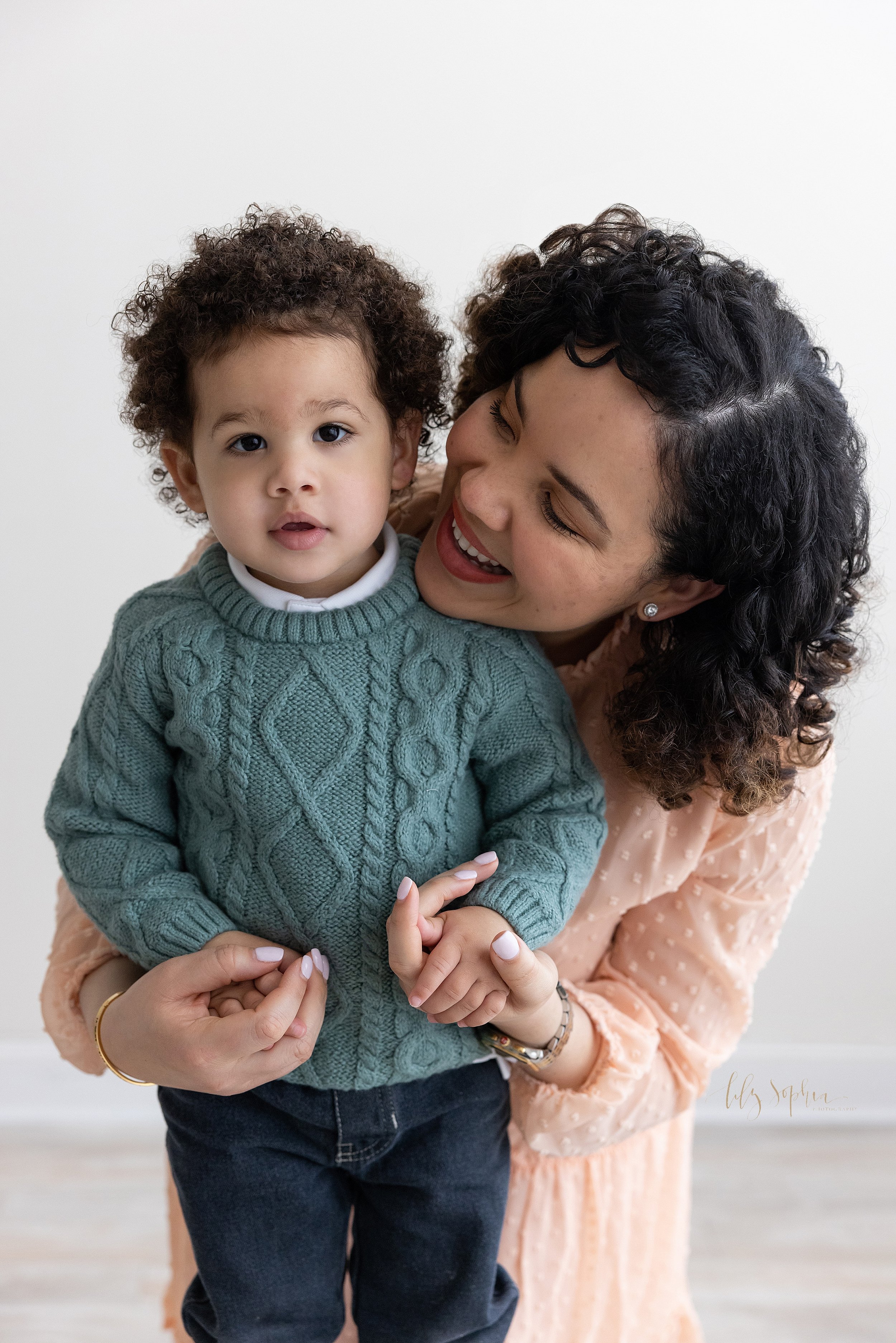  Family photo of a mother kneeling behind her two year old son as she holds his hands and peeks over his right shoulder to look at him taken near Kirkwood in Atlanta in a studio using natural light. 