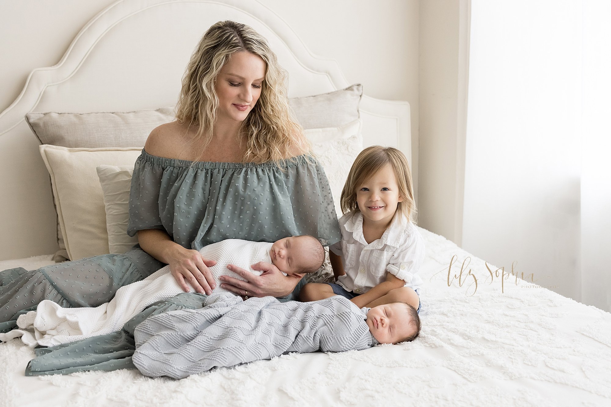  Newborn family portrait of a mother sitting on top of bed wearing an off the shoulder flocked gown holding her one twin son on her lap, her other twin son lies in front of her on the bed and her young daughter sits cross legged next to her with natu