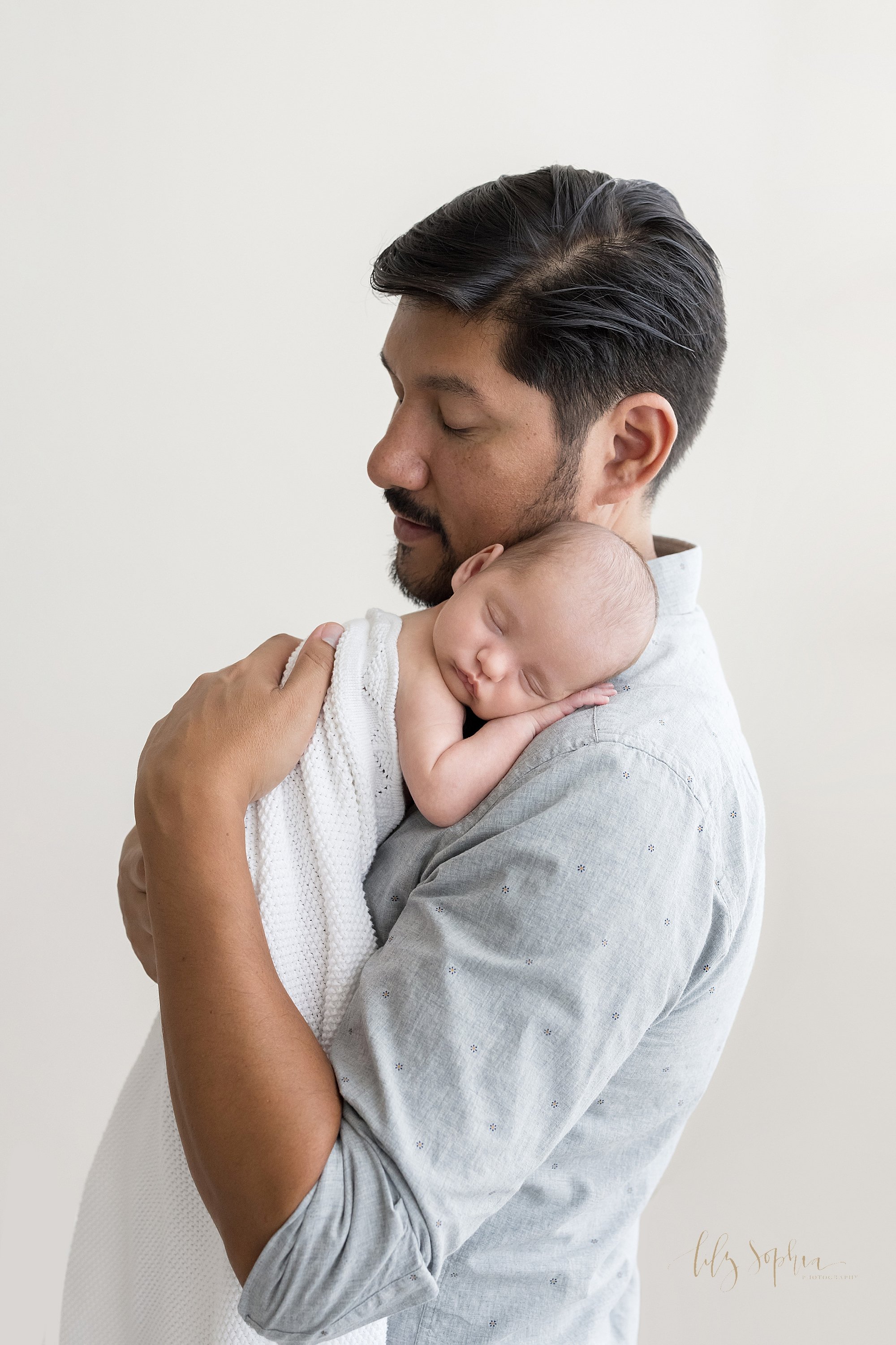  Newborn portrait of a father holding his infant son on his shoulder as he stands in front of a window streaming natural light cherishing this moment with his son taken near Ansley Park in Atlanta in a natural light studio. 