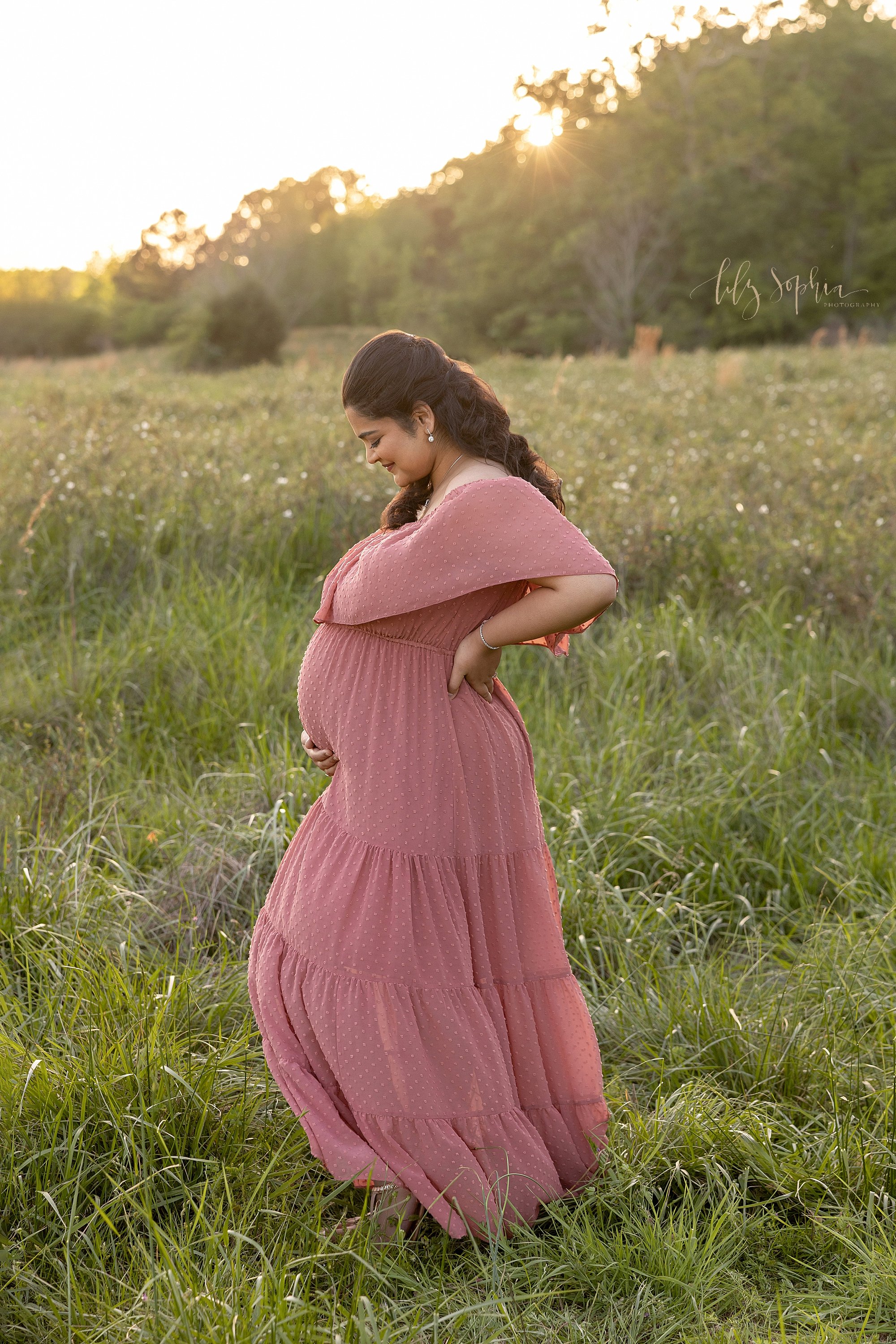  Maternity portrait of a pregnant mother standing facing the left to show her profile as she holds the base of her belly with her right hand and places her left hand on her hip while standing in at sunset in field near Atlanta. 