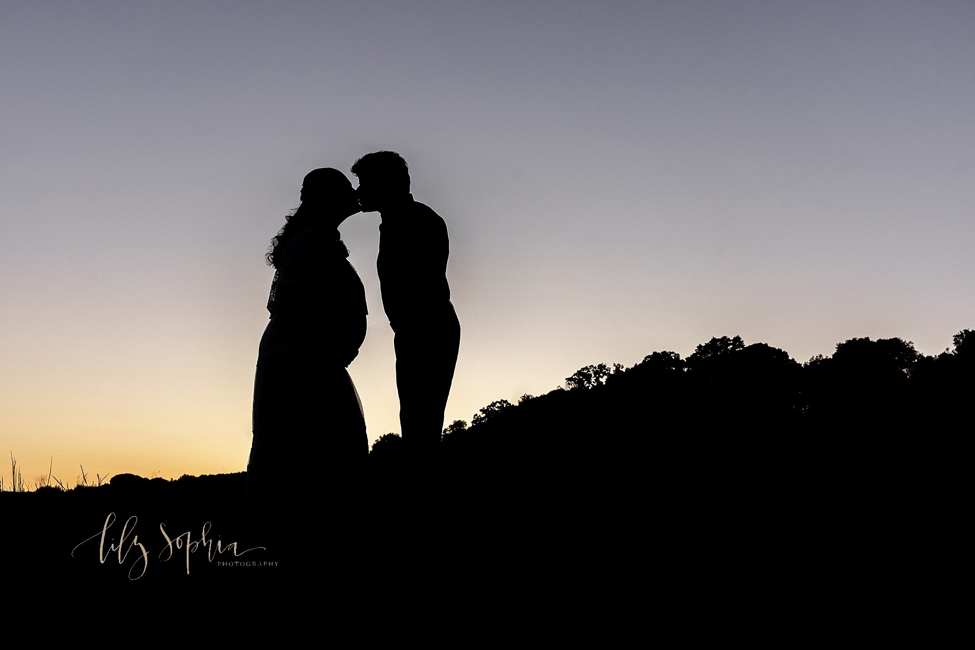  Silhouetted maternity portrait of an expectant couple as they stand on a ridge at sunset in a park near Atlanta.  