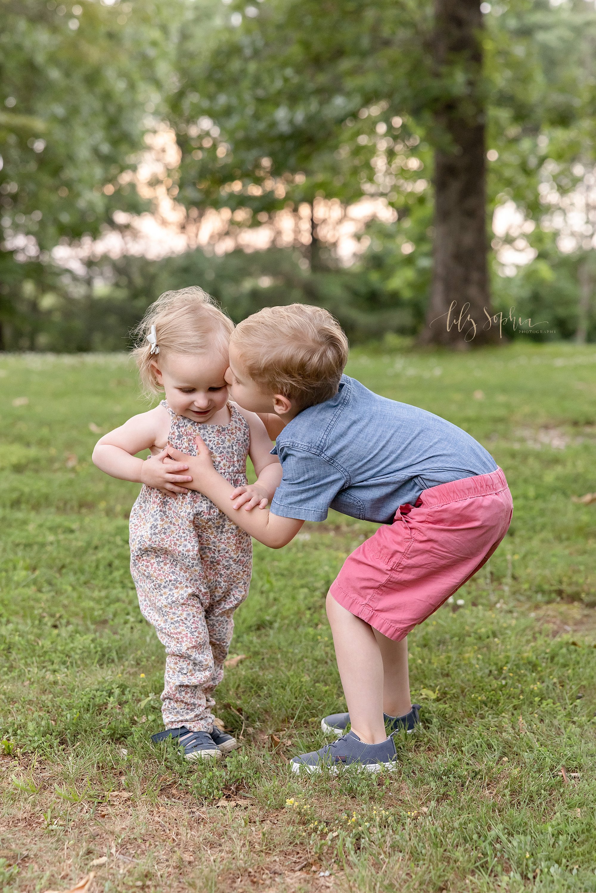  Family sibling photo of a toddler girl walking in a park as her older brother reaches down to give her a kiss taken near Atlanta at sunset. 