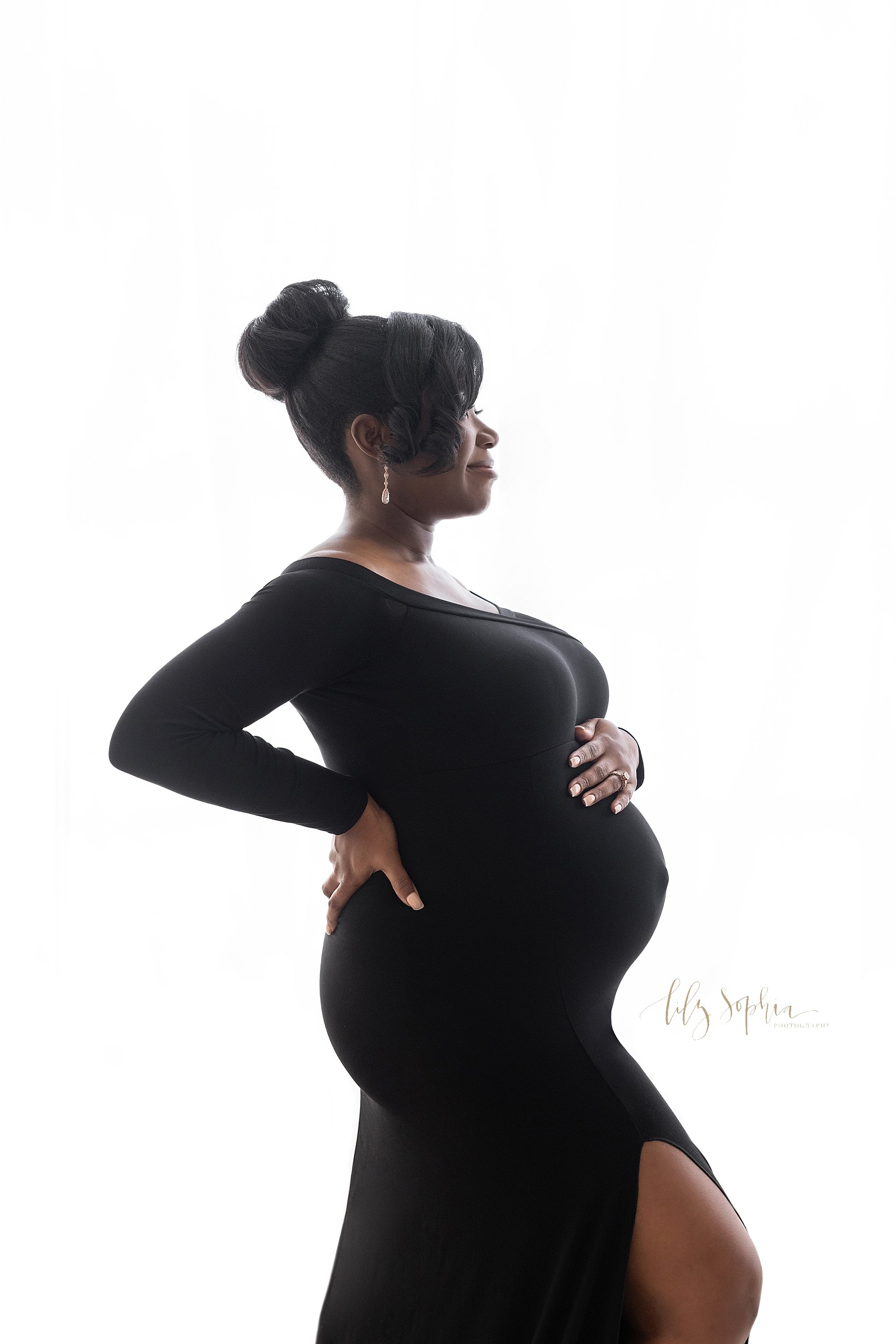  Maternity portrait of an African-American mother wearing a jersey knit long-sleeve gown with a side slit as she stands in front of a natural light window to show her profile with her left hand on top of her belly and her right hand on her hip taken 