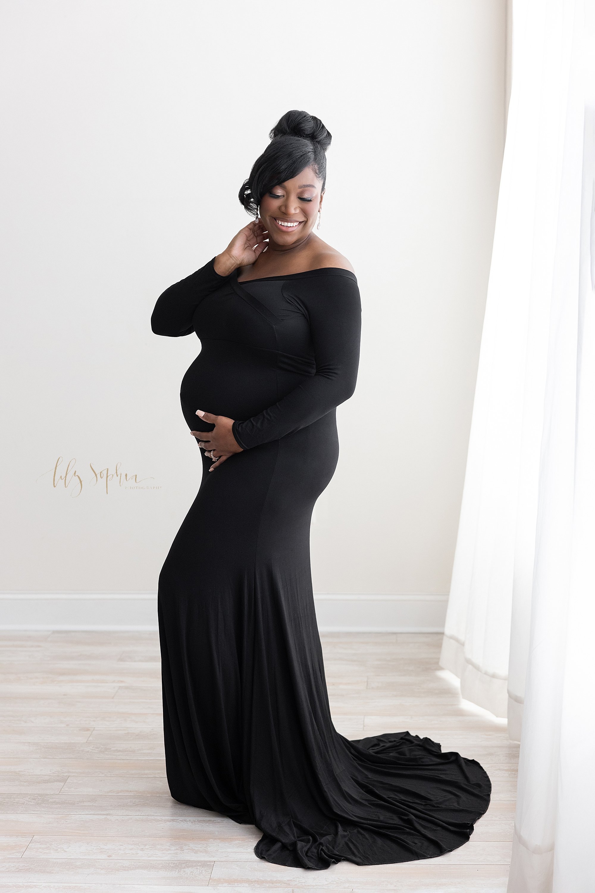  Maternity portrait of an African-American woman wearing a jersey knit long-sleeved gown with a train that flows on the floor behind her as she stands with her right hand at the base of her belly and her left hand touching her neck taken near Kirkwoo