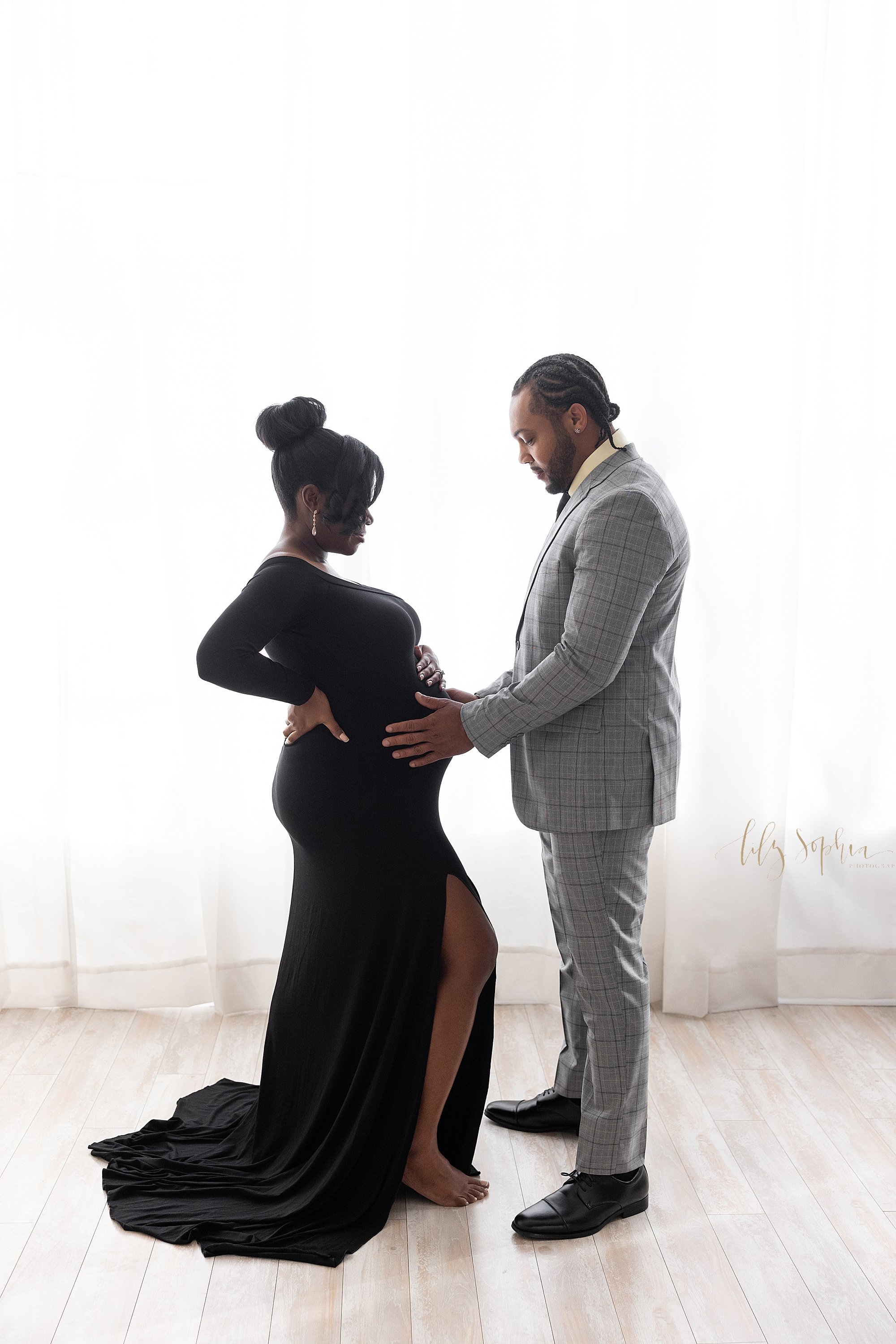  Maternity photo shoot of a pregnant African-American mother standing in front of a window streaming natural light with her right hand on her hip and her left hand on top of her belly as she faces her husband who has both hands on her belly and the t