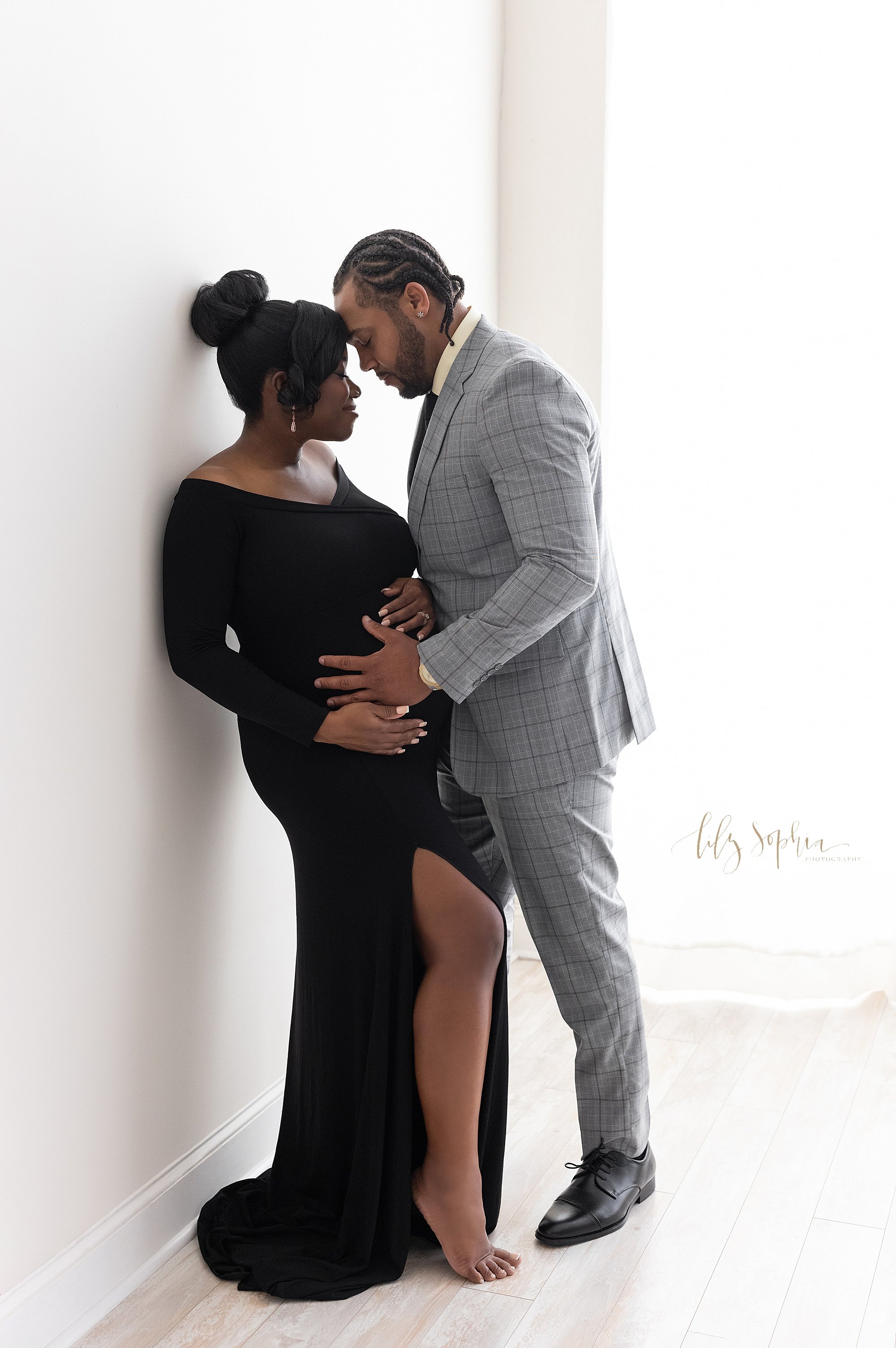  Maternity photo session of an African-American pregnant mother as she wears a sleek slit front knit long-sleeve gown and stands with her shoulders against a wall with her husband facing her wearing a plaid suit with his forehead touching his wife’s 