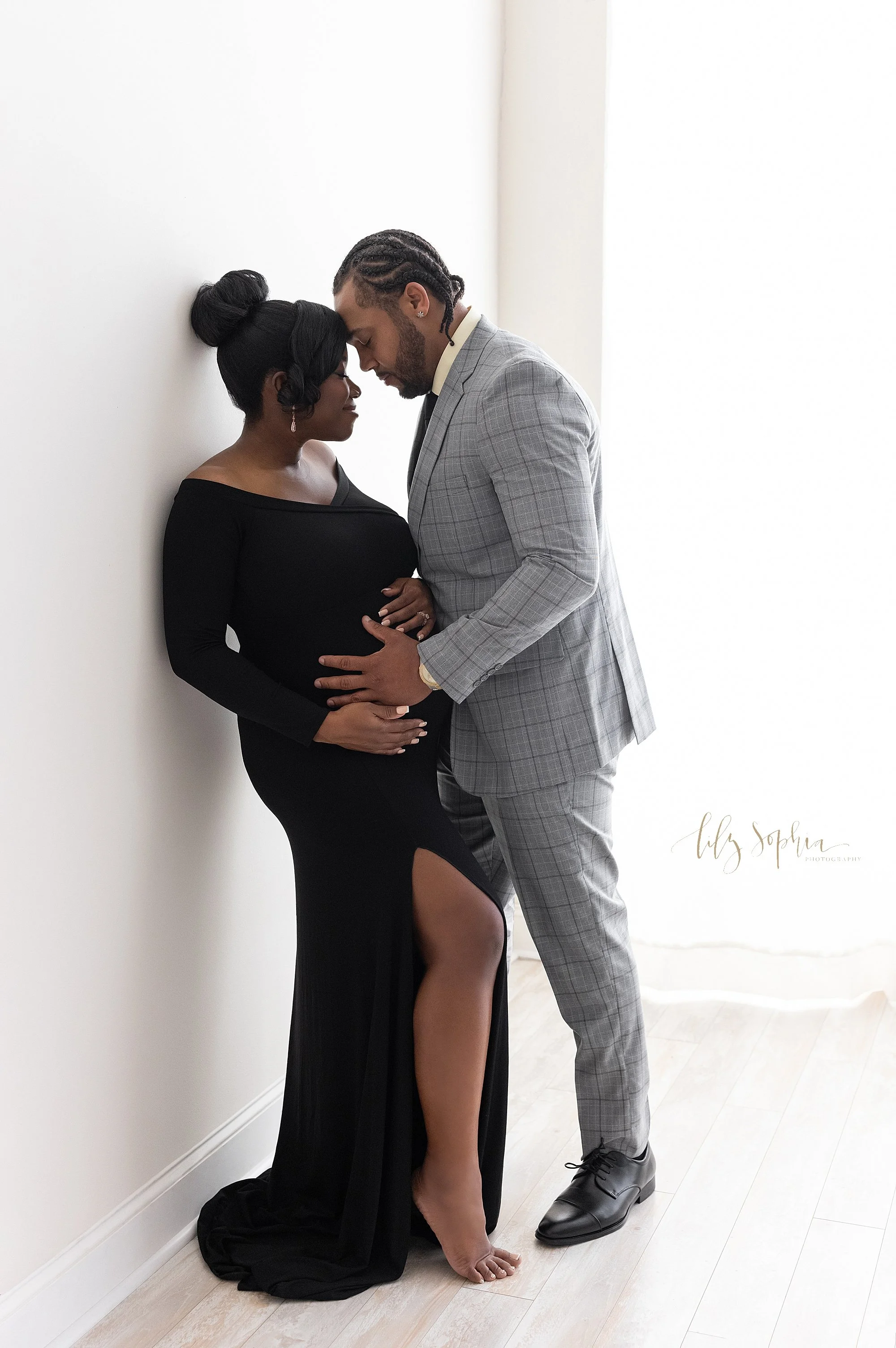  Maternity photo session of an African-American pregnant mother as she wears a sleek slit front knit long-sleeve gown and stands with her shoulders against a wall with her husband facing her wearing a plaid suit with his forehead touching his wife’s 