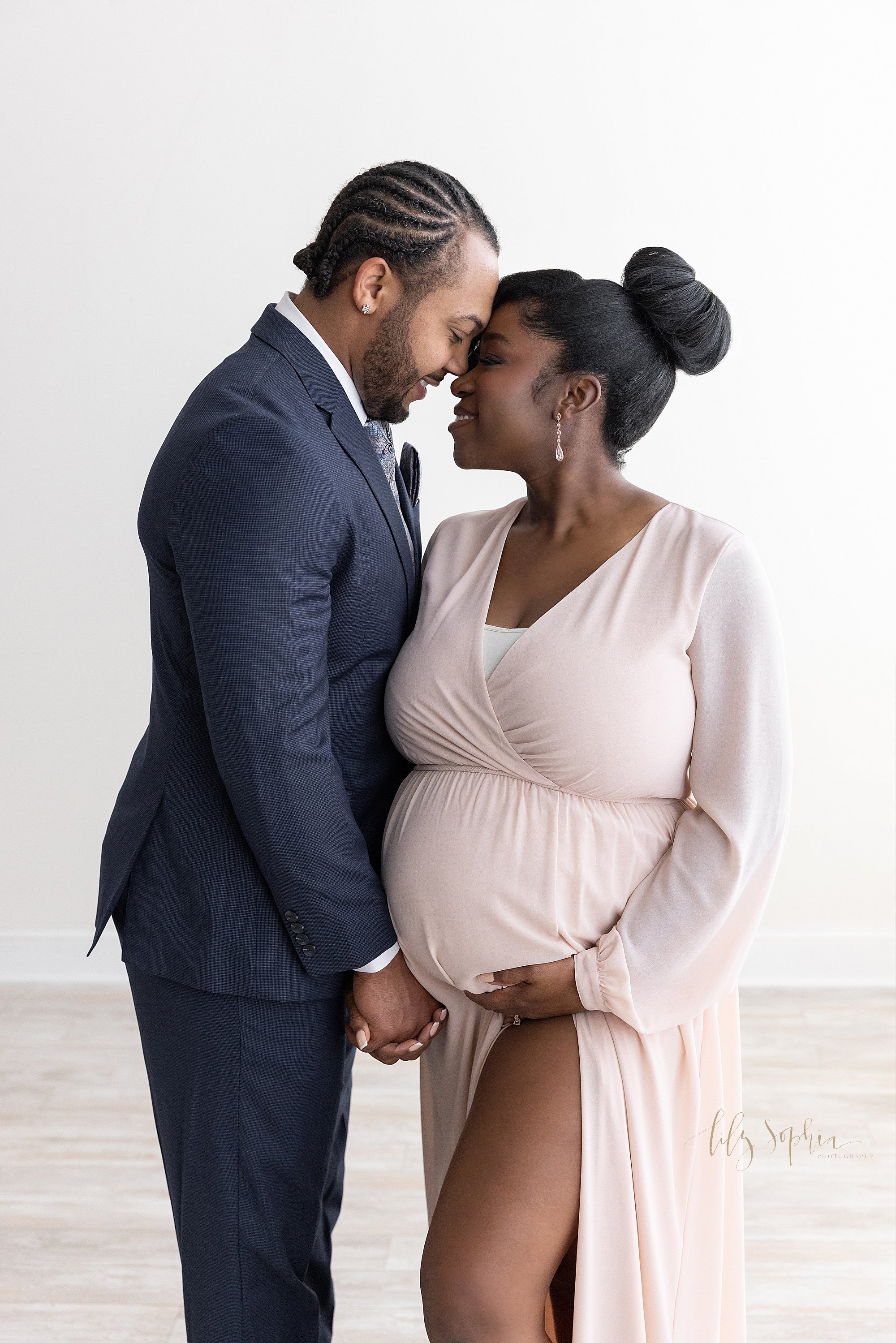  Maternity portrait of a pregnant African-American mother standing in a studio with her left hand holding her belly and her left leg peeking out from her flowing knit gown and she and her husband hold hands and her bends to kiss her taken near Virgin
