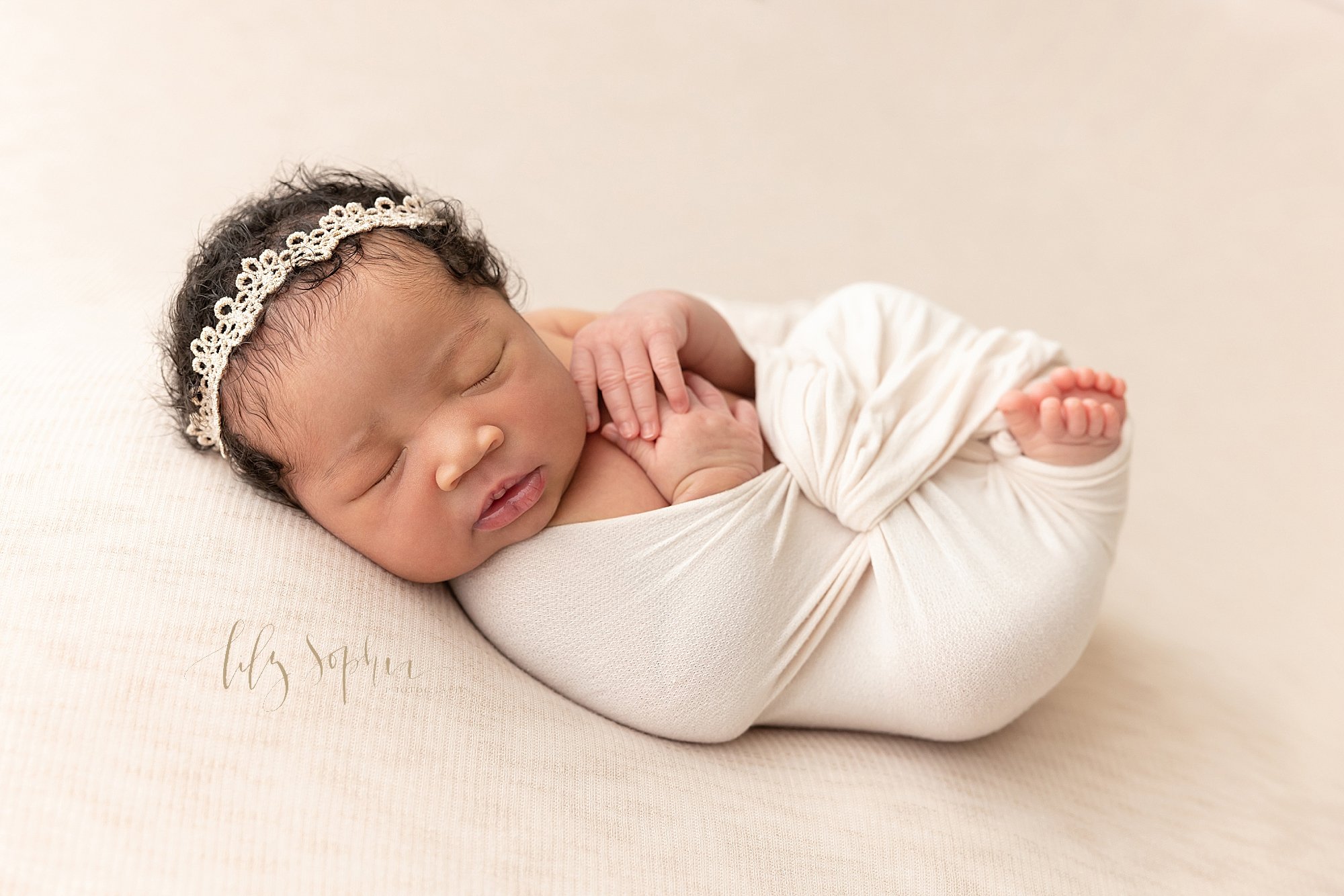  Newborn photo of a peacefully sleeping bundle of joy as an African-American baby girl wearing a delicate crocheted headband on her curly black head lies on her back bundled in a stretchy swaddle with her toes peeking out taken in a photography studi