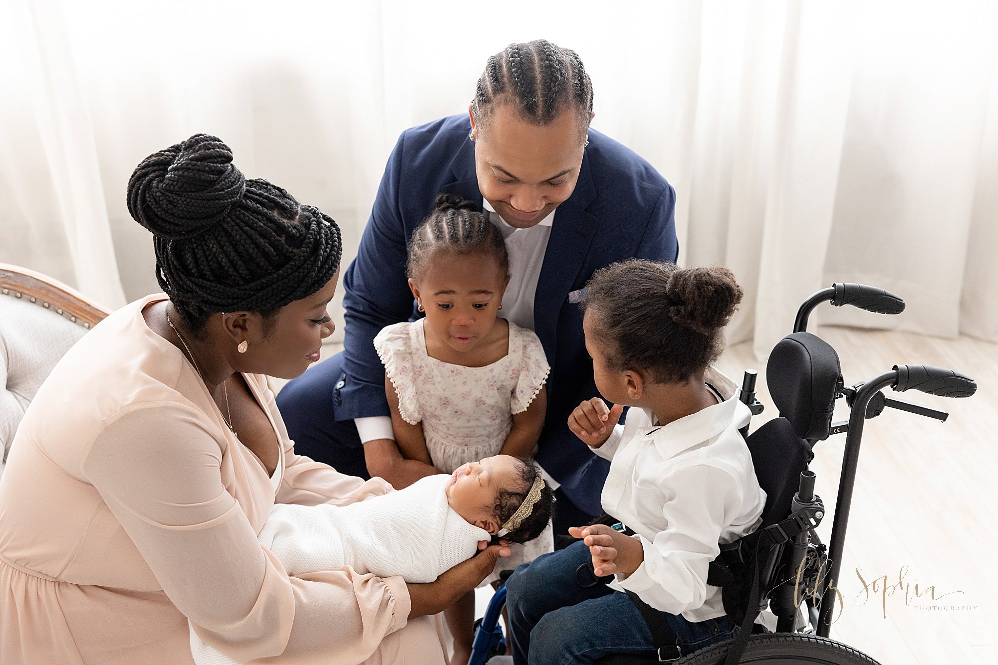  Family newborn photo shoot of an African-American mother sitting on an  tufted sofa holding her newborn daughter on her lap with her husband squatting on her left side with their toddler daughter in front of him and their special needs son sitting i