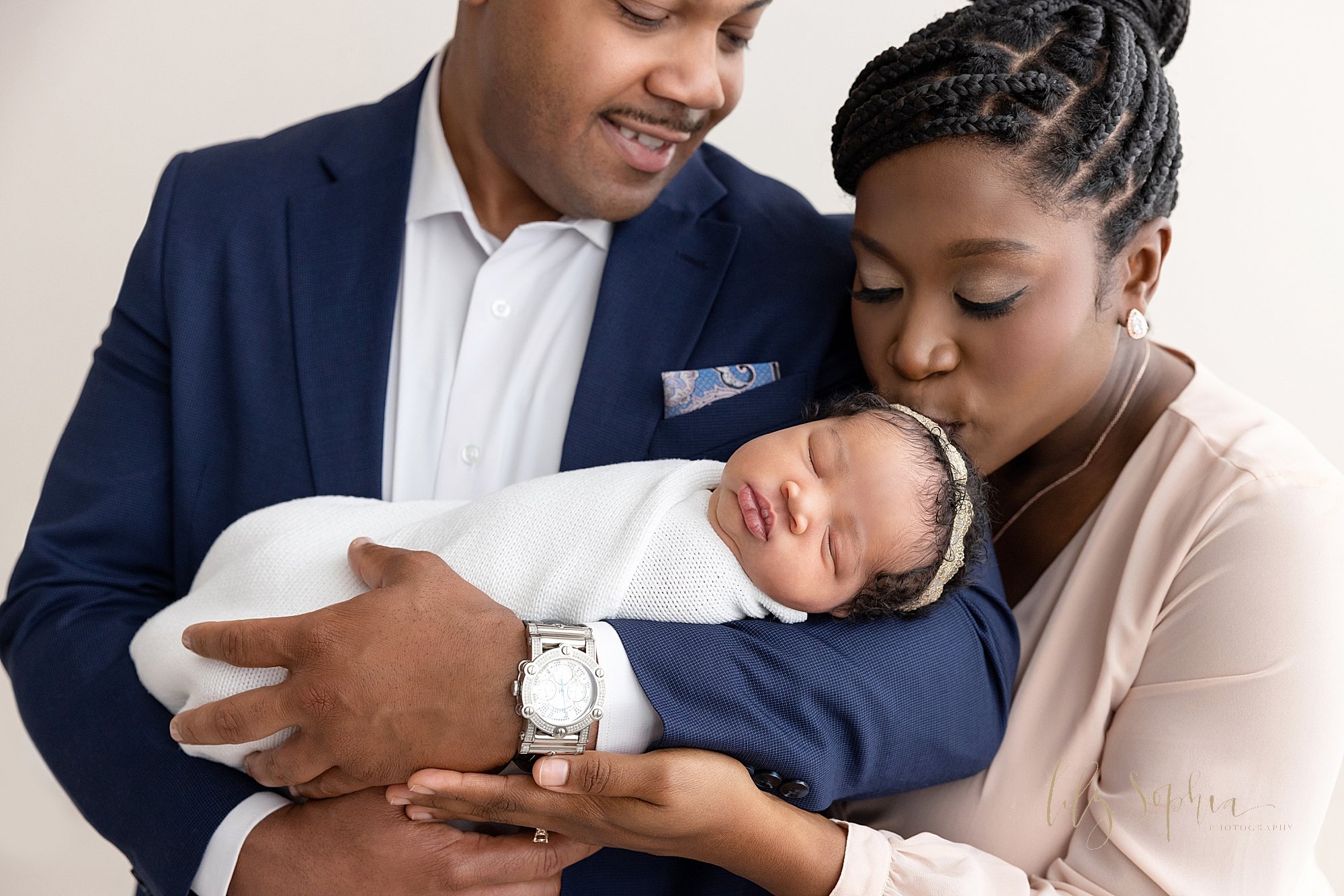  Newborn photo session with an African-American father cradling his newborn baby daughter in his arms as mom stands to his left and leans down to kiss the crown of her sleeping newborn daughter’s head taken in a natural light studio near Decatur in A