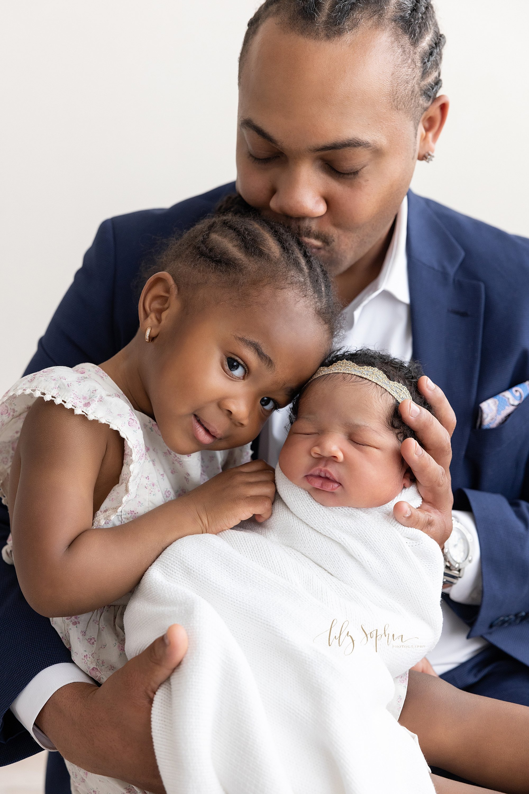  Newborn portrait of an African-American father holding his newborn daughter in front of him as he sits with his toddler daughter on his right knee with the toddler placing her forehead next to her sister’s head and her father kissing the top of her 