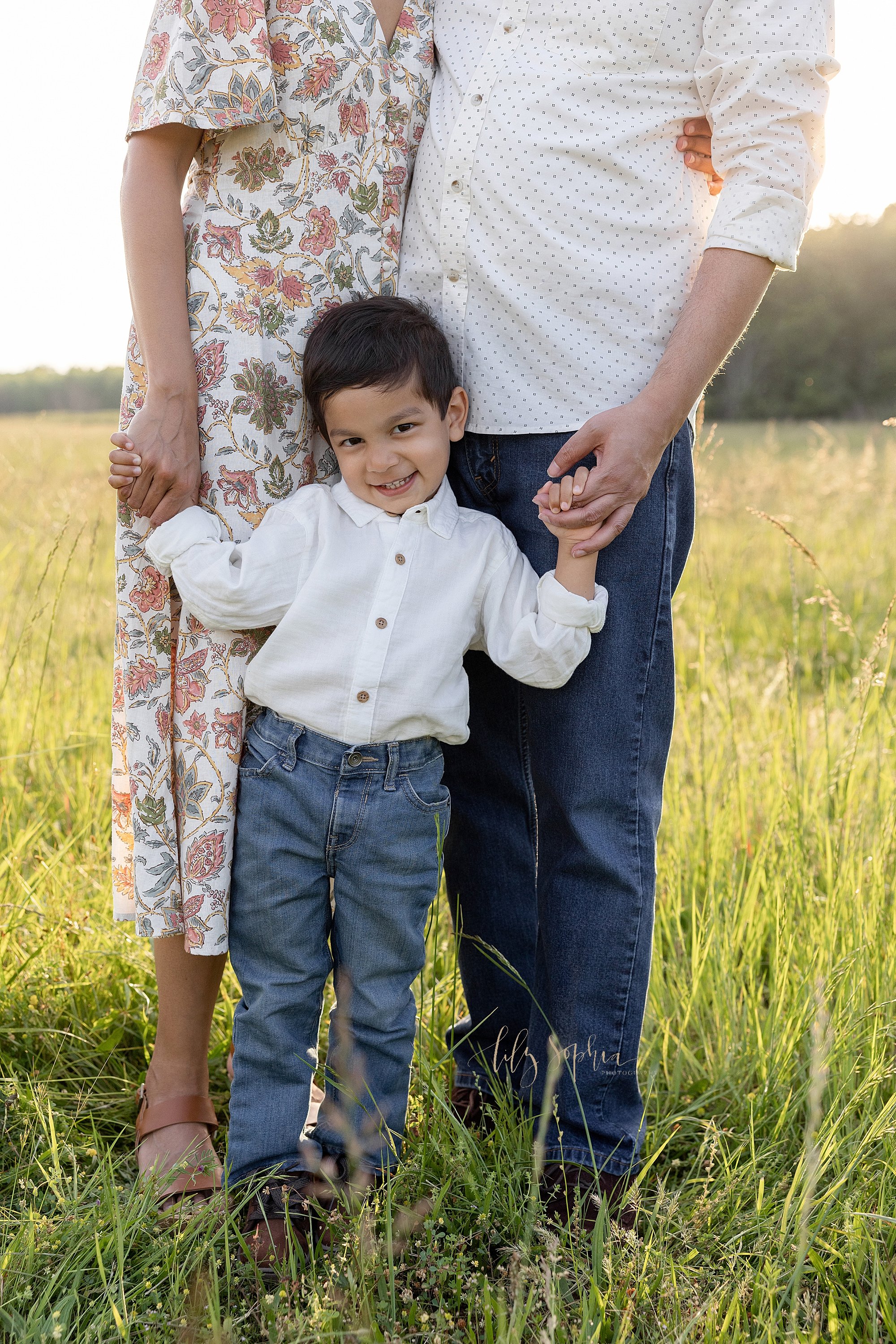  Family photo of a toddler boy standing in front of his mother and father as he holds each of their hands at sunset in an Atlanta, Georgia field. 
