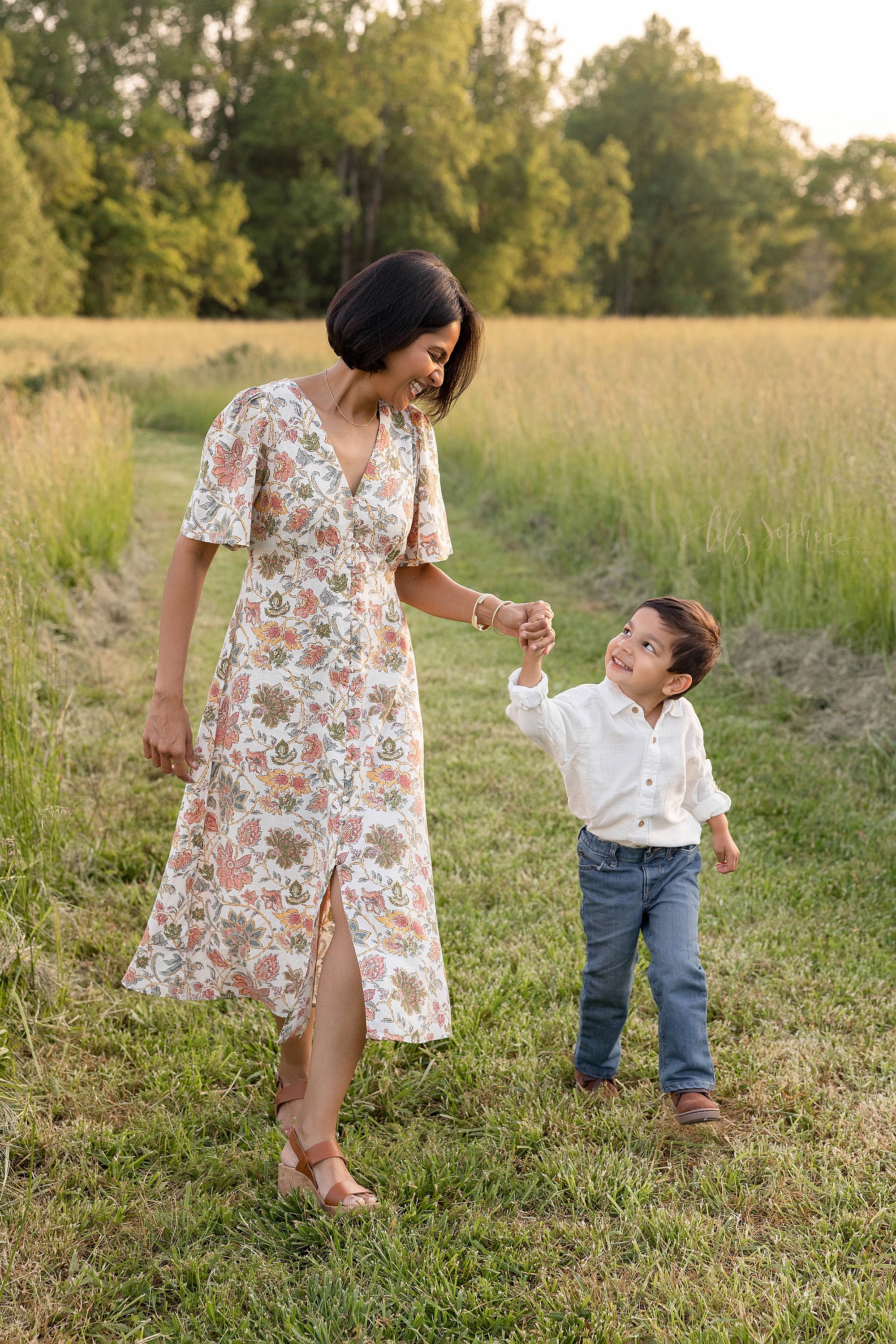  Family photograph with a mother and son walking hand in hand down a mown path in an Atlanta field at sunset as the little boy looks up to his mom and she smiles at him. 