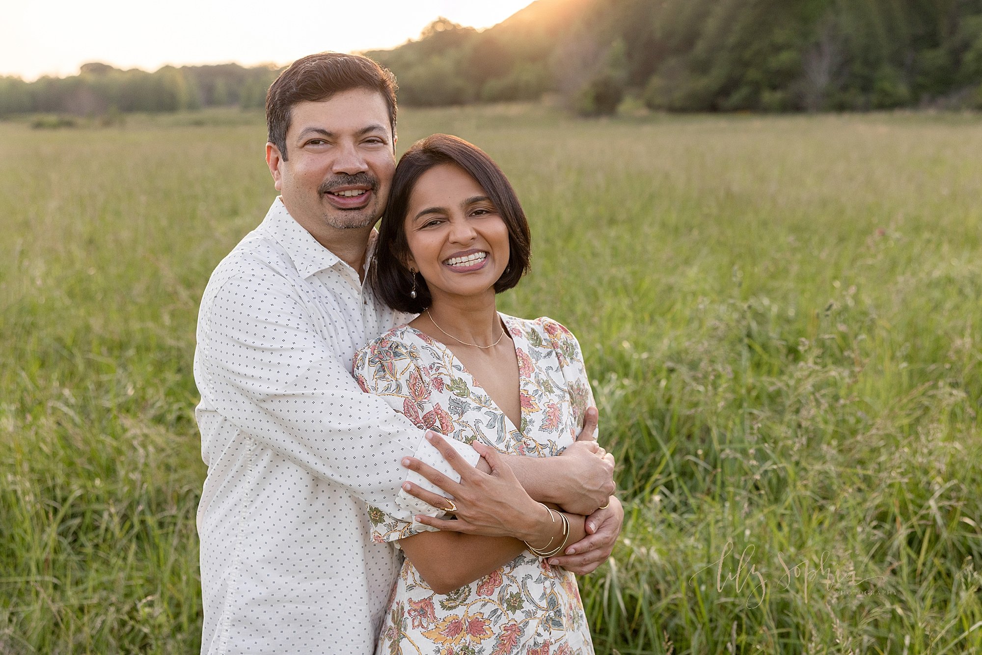 Family picture of a husband standing behind his wife as he wraps his arms around her with the sun setting in the background taken in Atlanta in a field. 