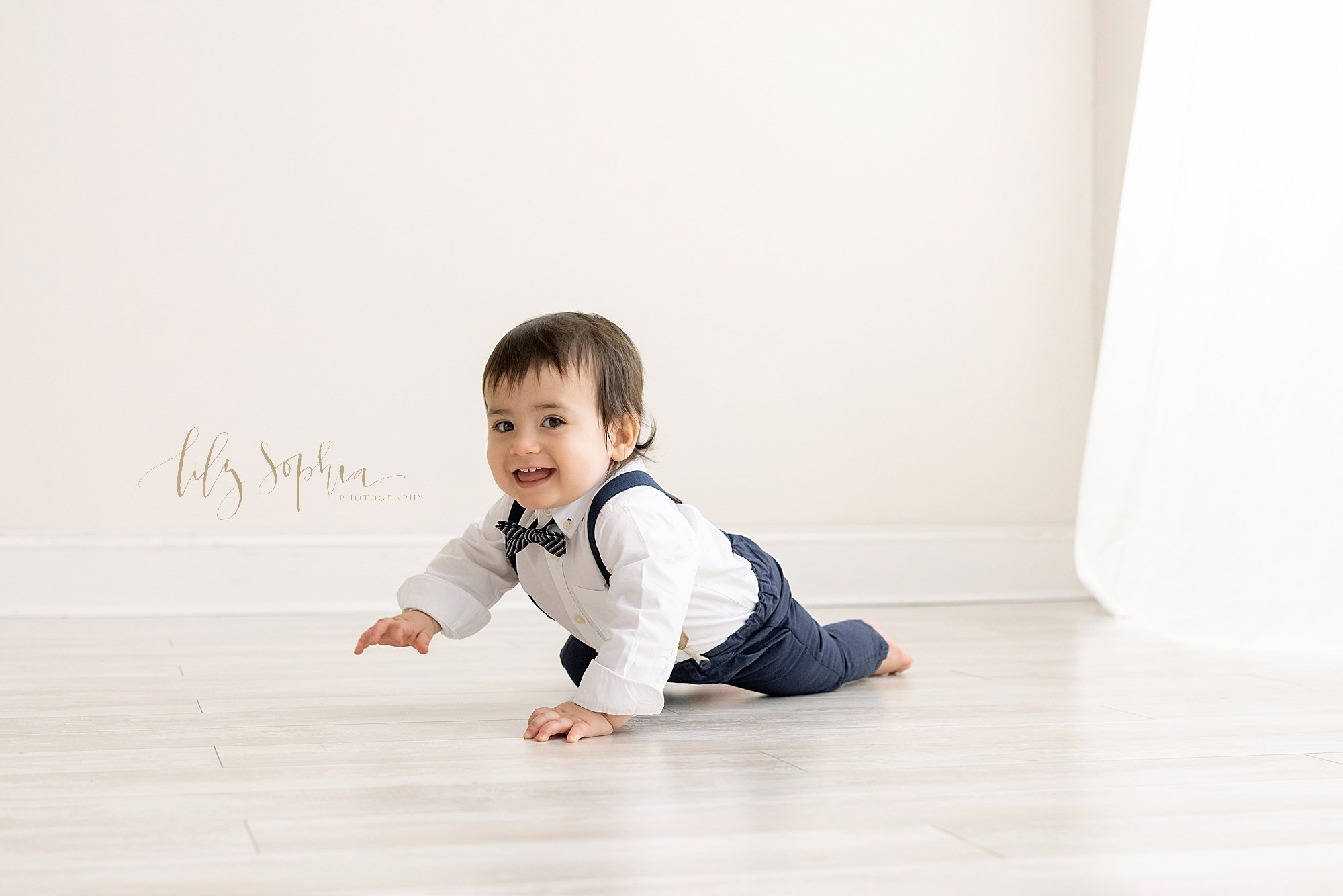  First birthday photo of a one year old little boy as he crawls across the floor wearing a white shirt, blue bow tie, blue suspenders, and blue pants taken in front of a window streaming natural light in a photography studio in Ponce City Market in A