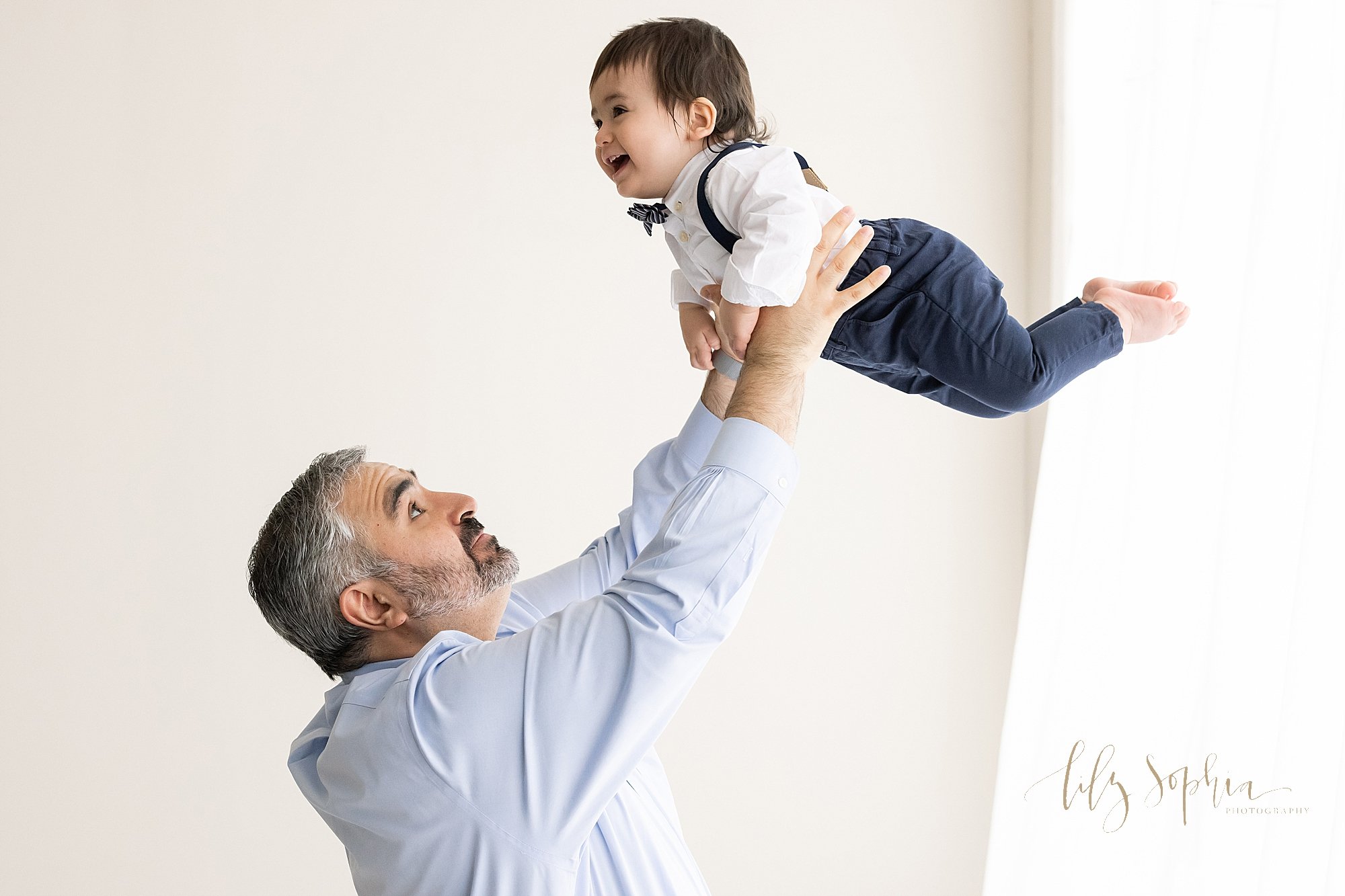  First birthday photo of a father throwing his one year old son above his head as his son squeals with delight taken in a natural light studio near Buckhead in Atlanta. 