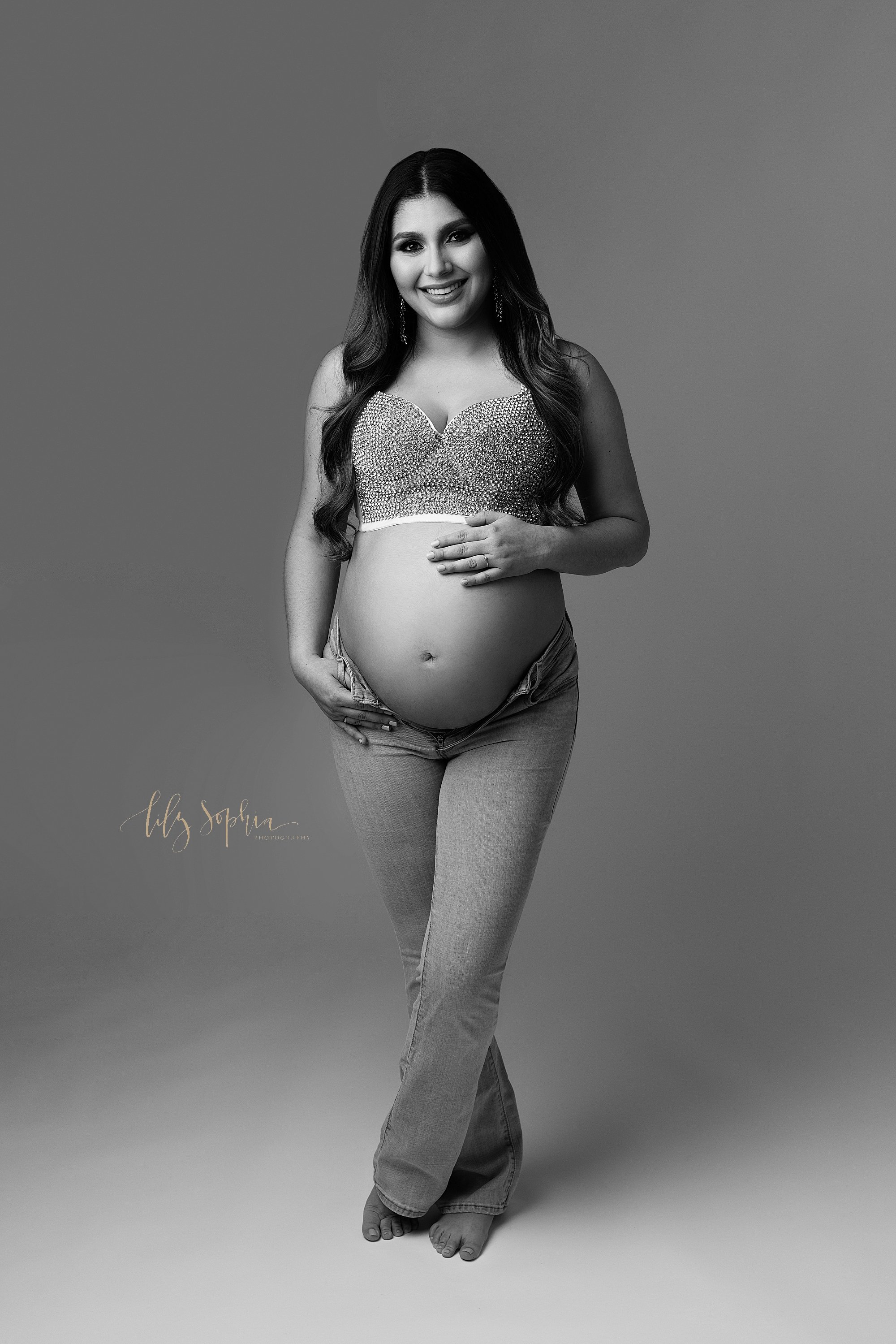  Modern maternity photo of a pregnant mother wearing a beaded bra and a pair of unzipped blue jeans with her right thumb hooked into the belt loop of her jeans and her left hand resting on her bare belly taken near Midtown in Atlanta in a natural lig