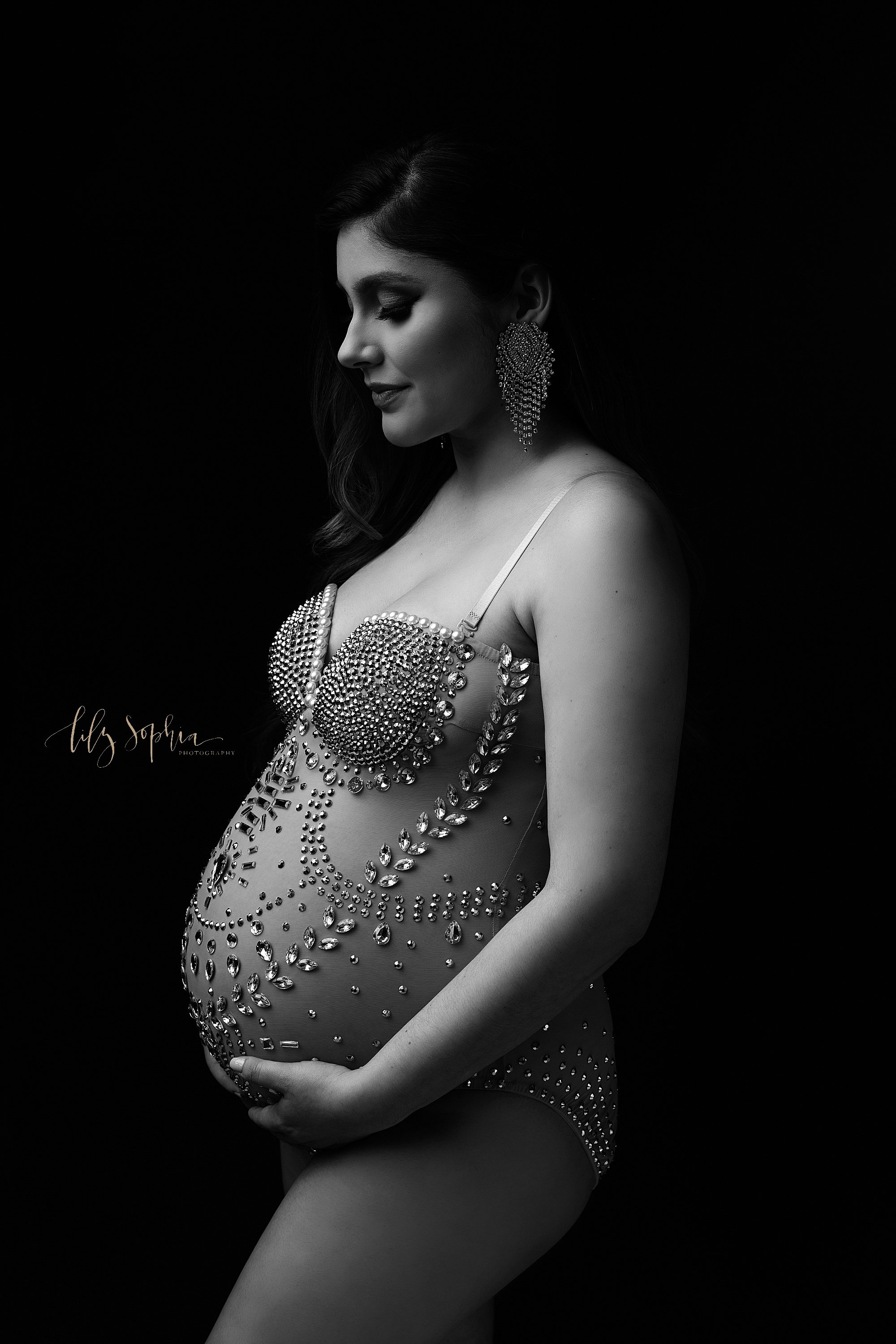  Modern maternity photo of a pregnant mother wearing a sheer one piece bejeweled bodysuit as the woman faces the left, holds the base of her belly, and contemplates the upcoming birth of her child taken using natural light  near Kirkwood in Atlanta i