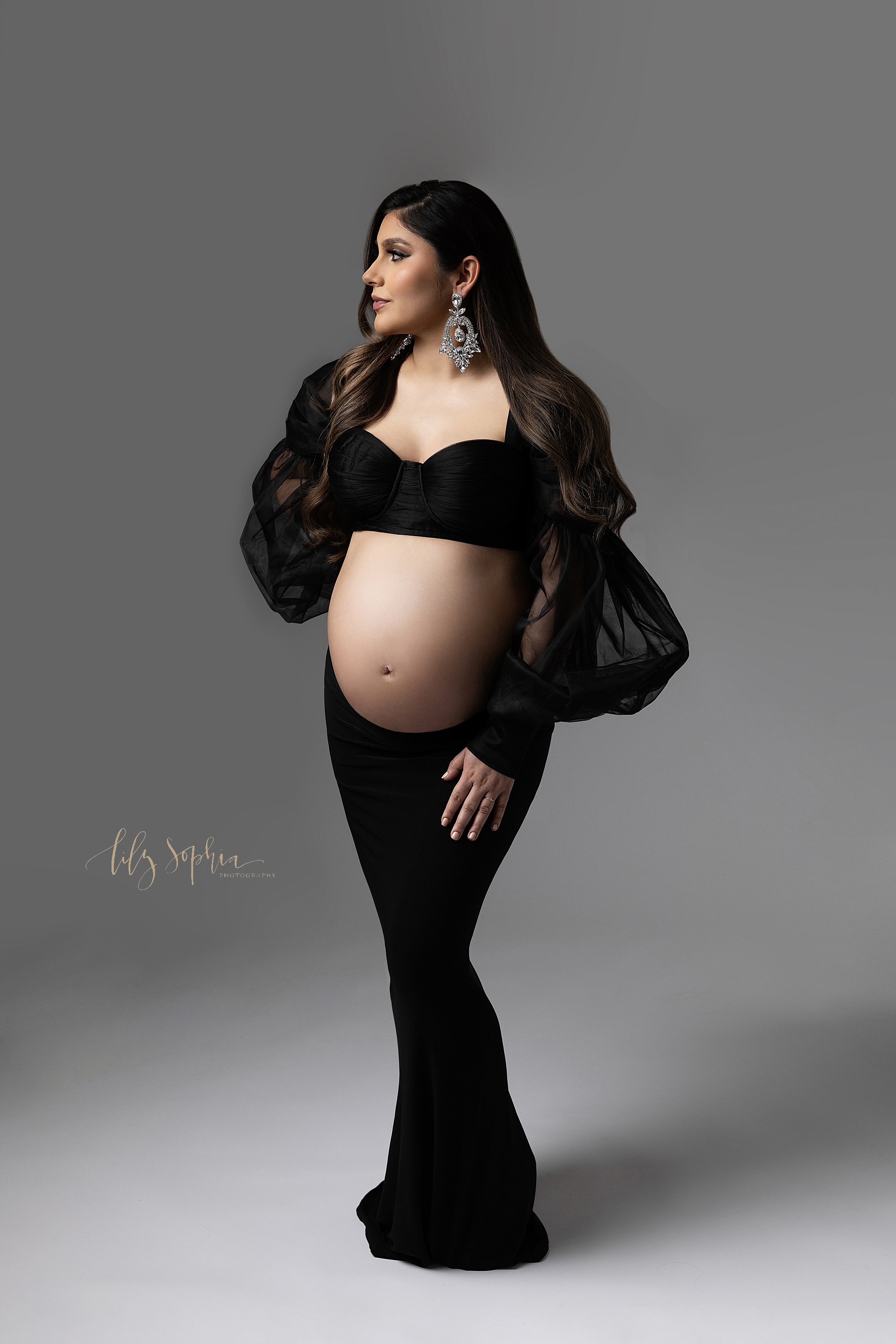  Modern maternity photo session with a pregnant mother wearing a black bra with a sheer black shrug and a full-length jersey knit skirt with her bare belly showing as she turns her head to look over her right shoulder taken in a natural light studio 
