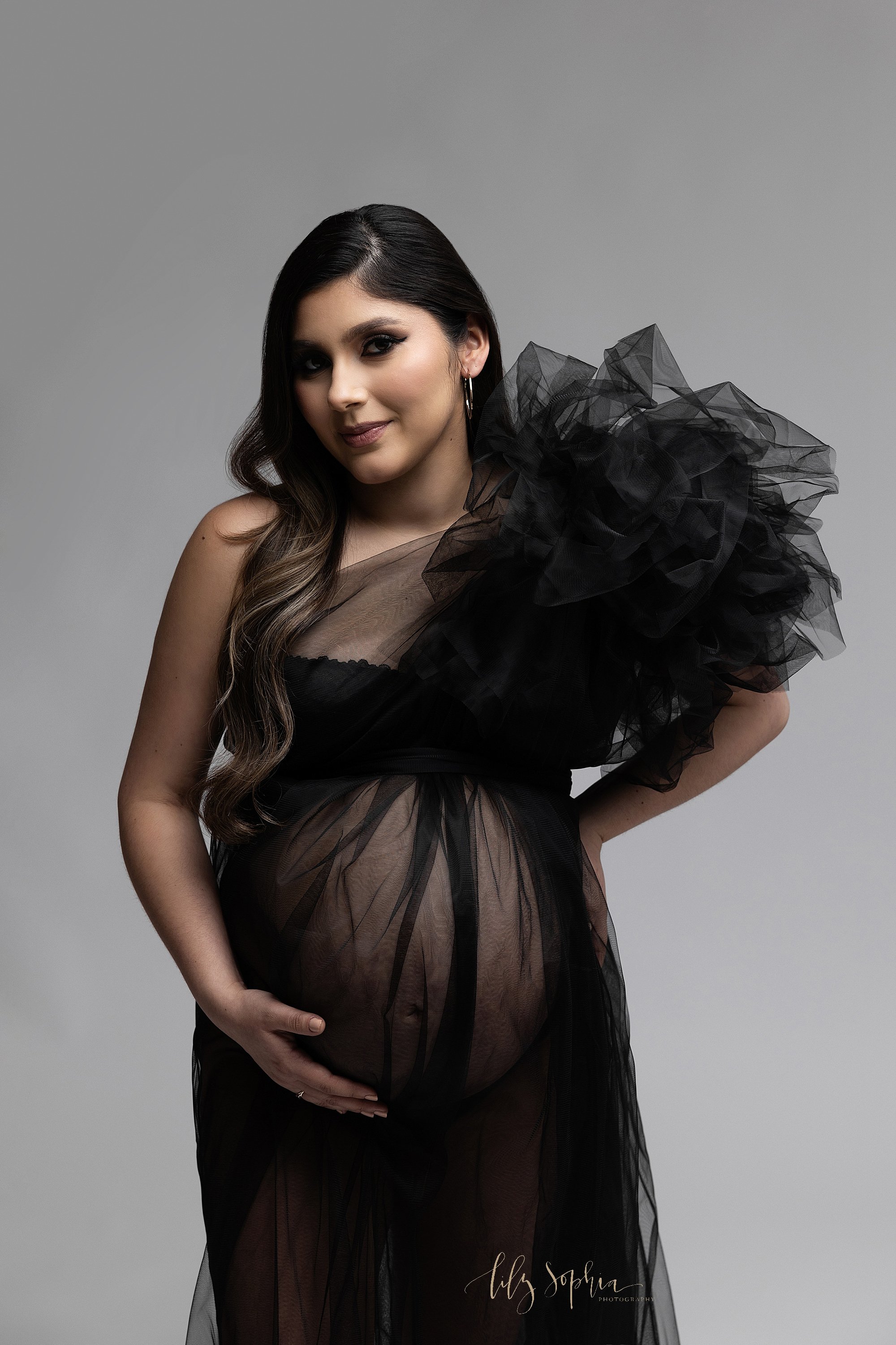  Modern maternity photo shoot with a pregnant mother wearing a black bandeau bra covered by a sheer full length gown with a large tulle flower on her left shoulder as she stands with her right hand holding the base of her belly and her left hand on h
