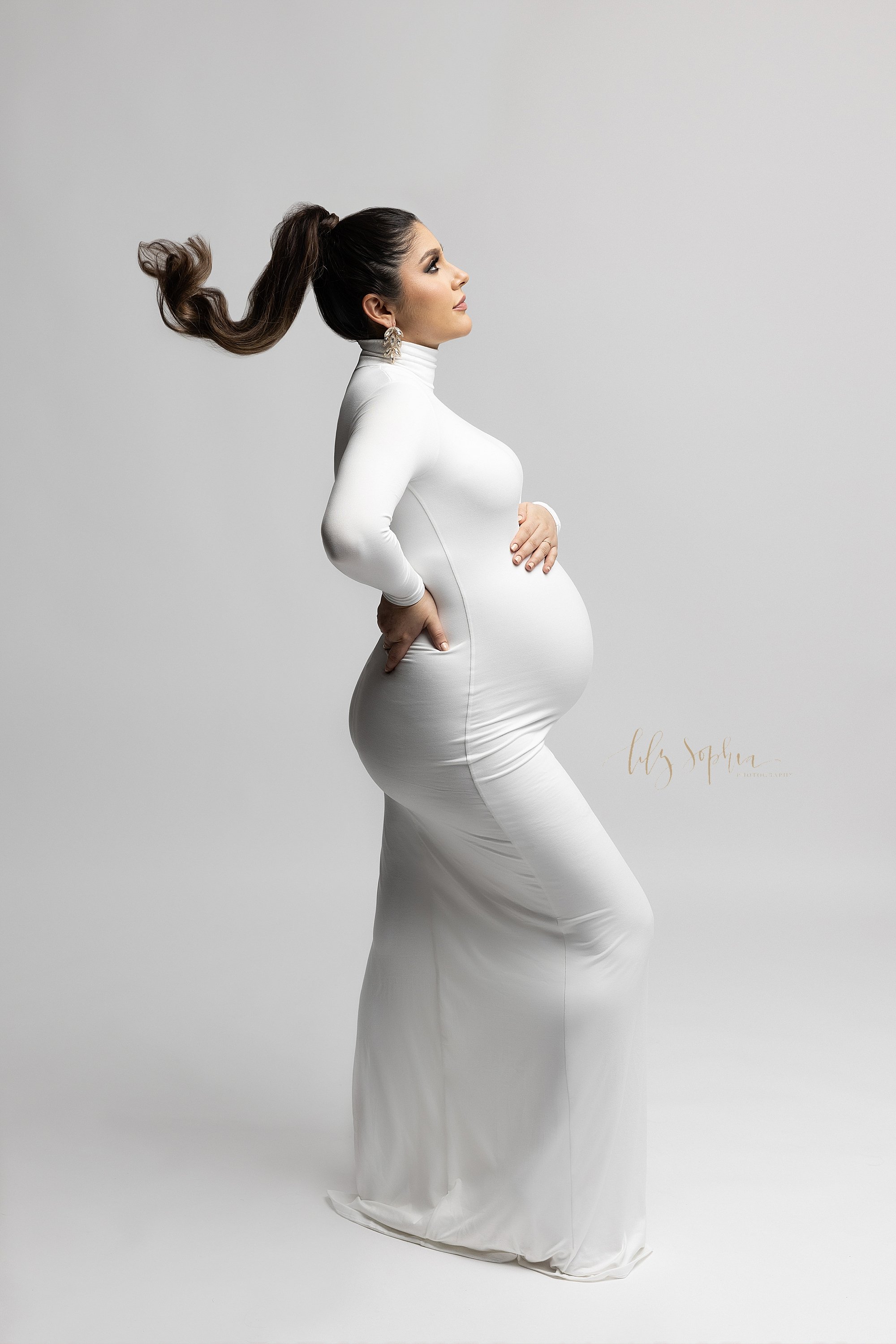 Modern maternity portrait  of a pregnant mother with a pony tail in her hair as it flies behind her while she stands facing the right wearing a jersey knit turtleneck one piece full-length dress with long sleeves as she places her right hand on top 