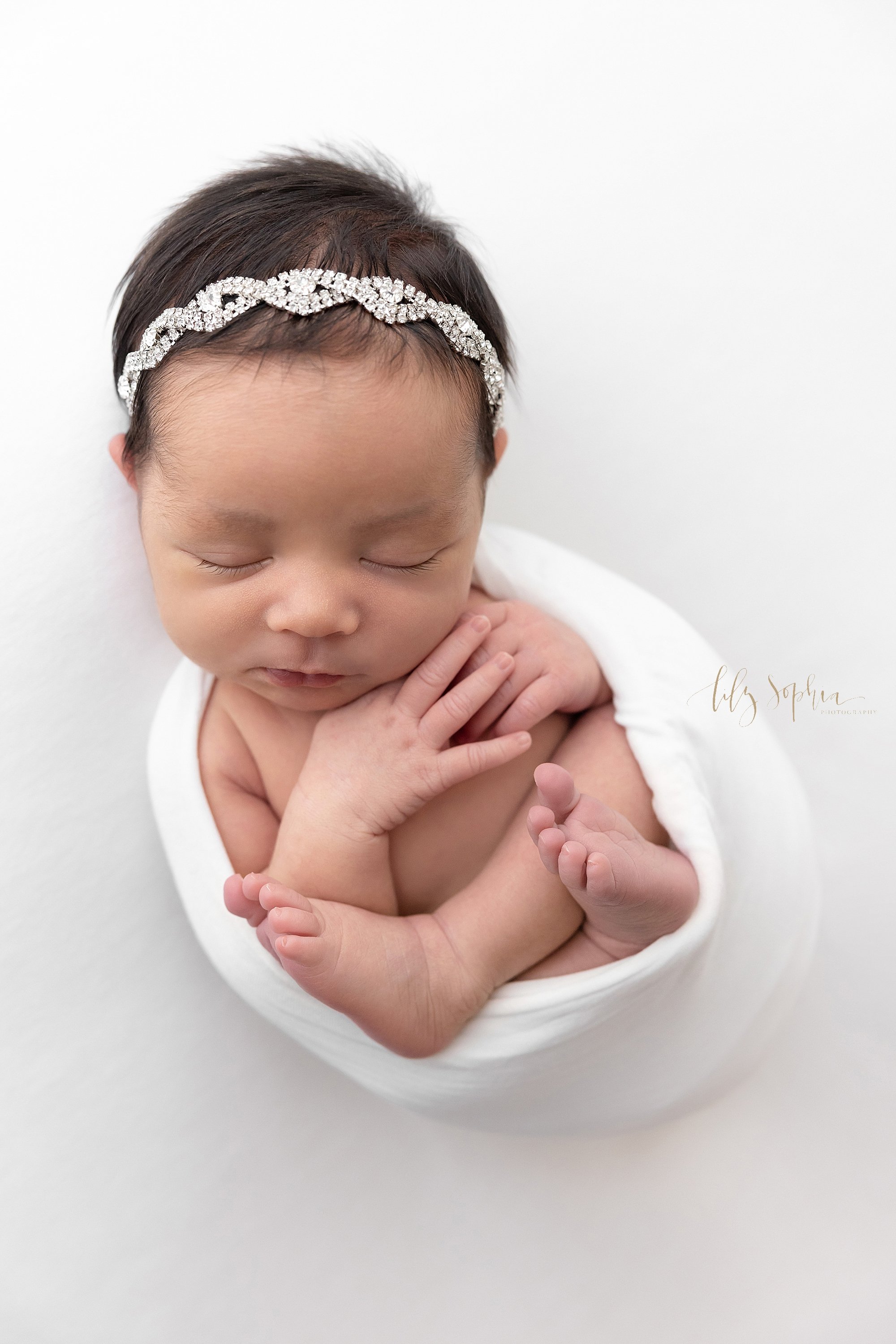  Newborn photo of a newborn baby girl wearing a rhinestone headband in her black wispy hair as she is cradled in a stretchy swaddle taken using natural light near Alpharetta in Atlanta in a photography studio. 