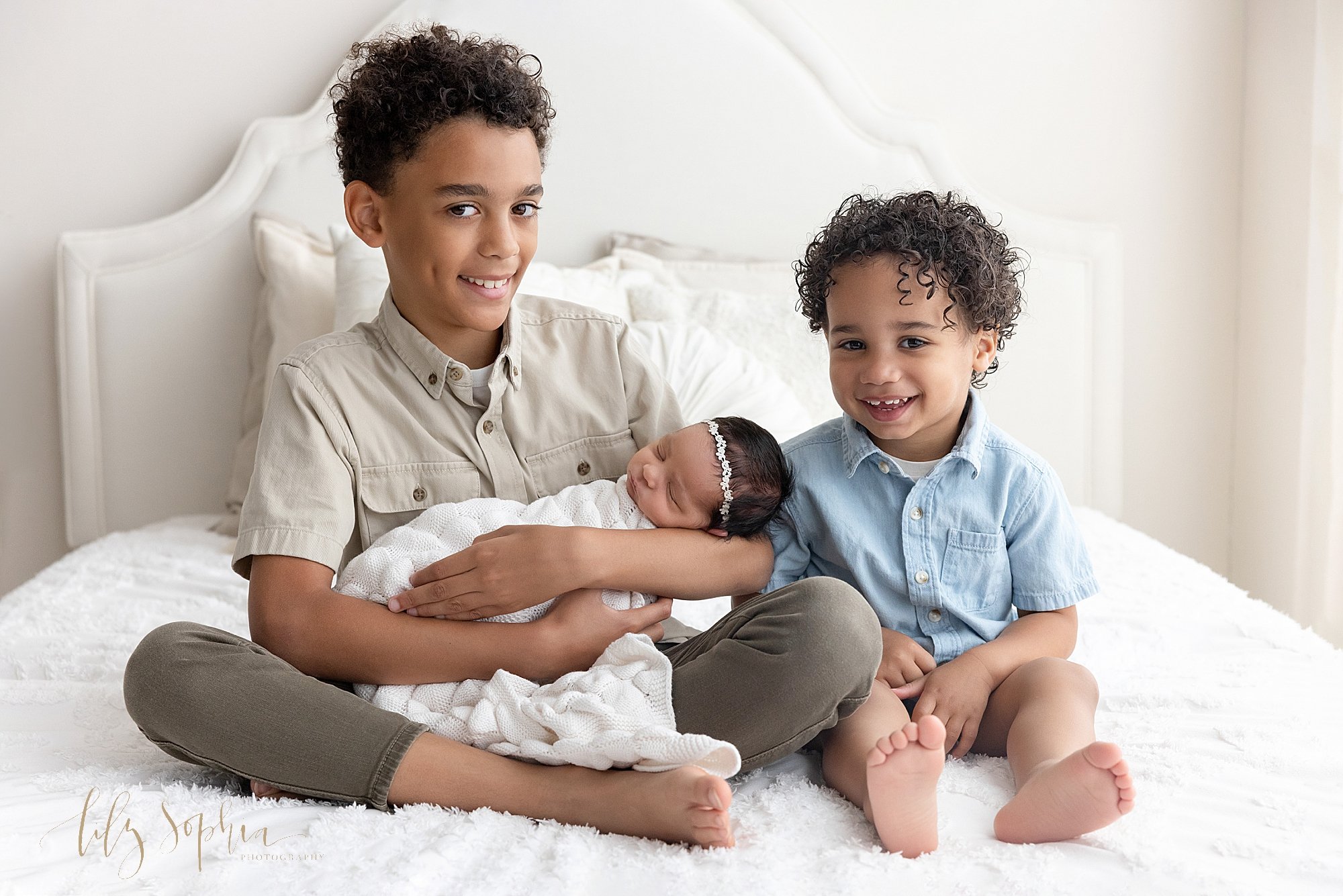  Family sibling portrait of a young boy cradling his newborn baby sister in is arms as he sits cross legged on a bed next to his toddler brother taken near Poncey Highlands in Atlanta in front of a window streaming natural light in a photography stud