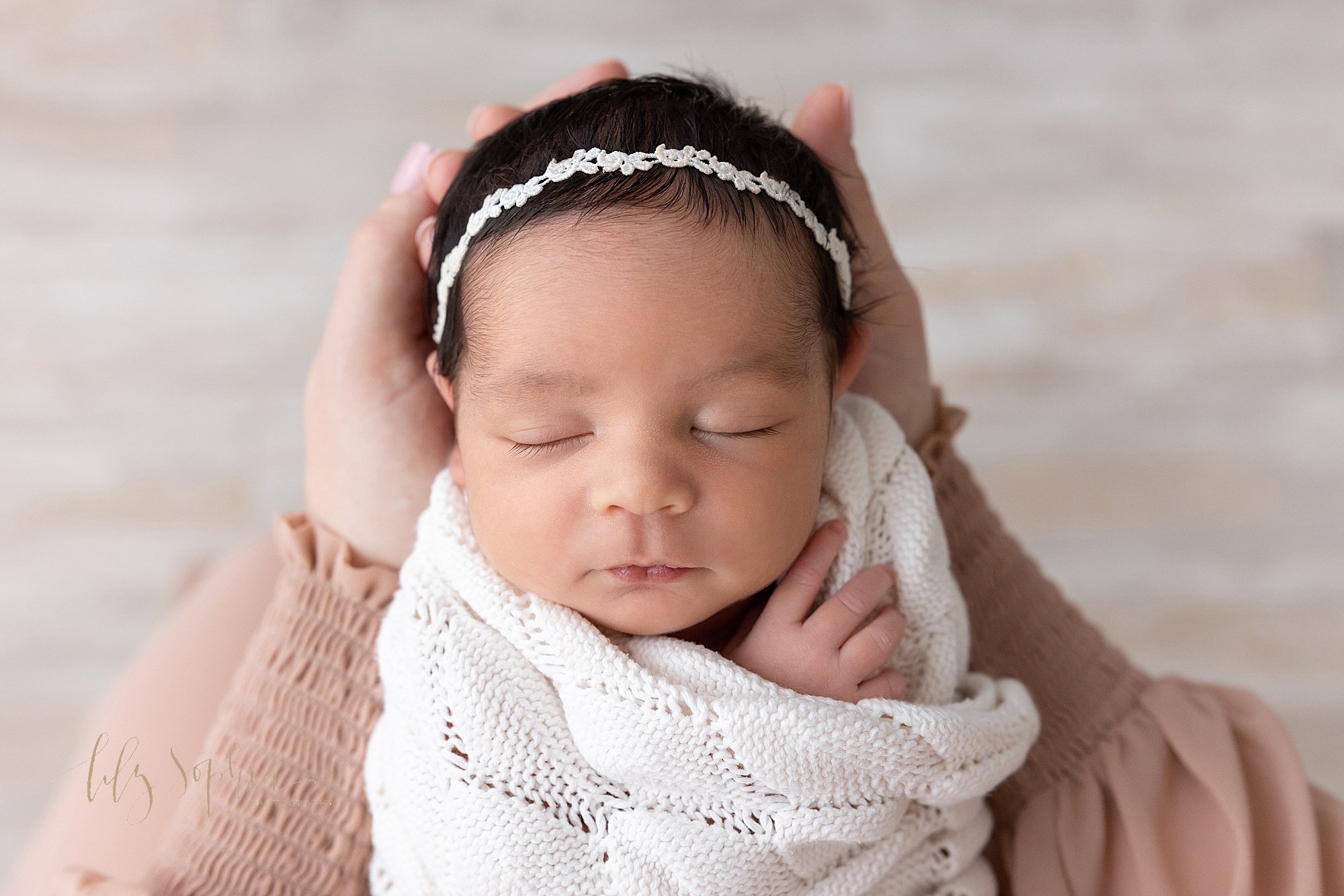  Cloe-up newborn portrait of a newborn baby girl wrapped in a soft white knitted blanket and wearing a delicate headband in her black hair as she peacefully sleeps while her head is held in her mother’s hands taken near Decatur in Atlanta using natur