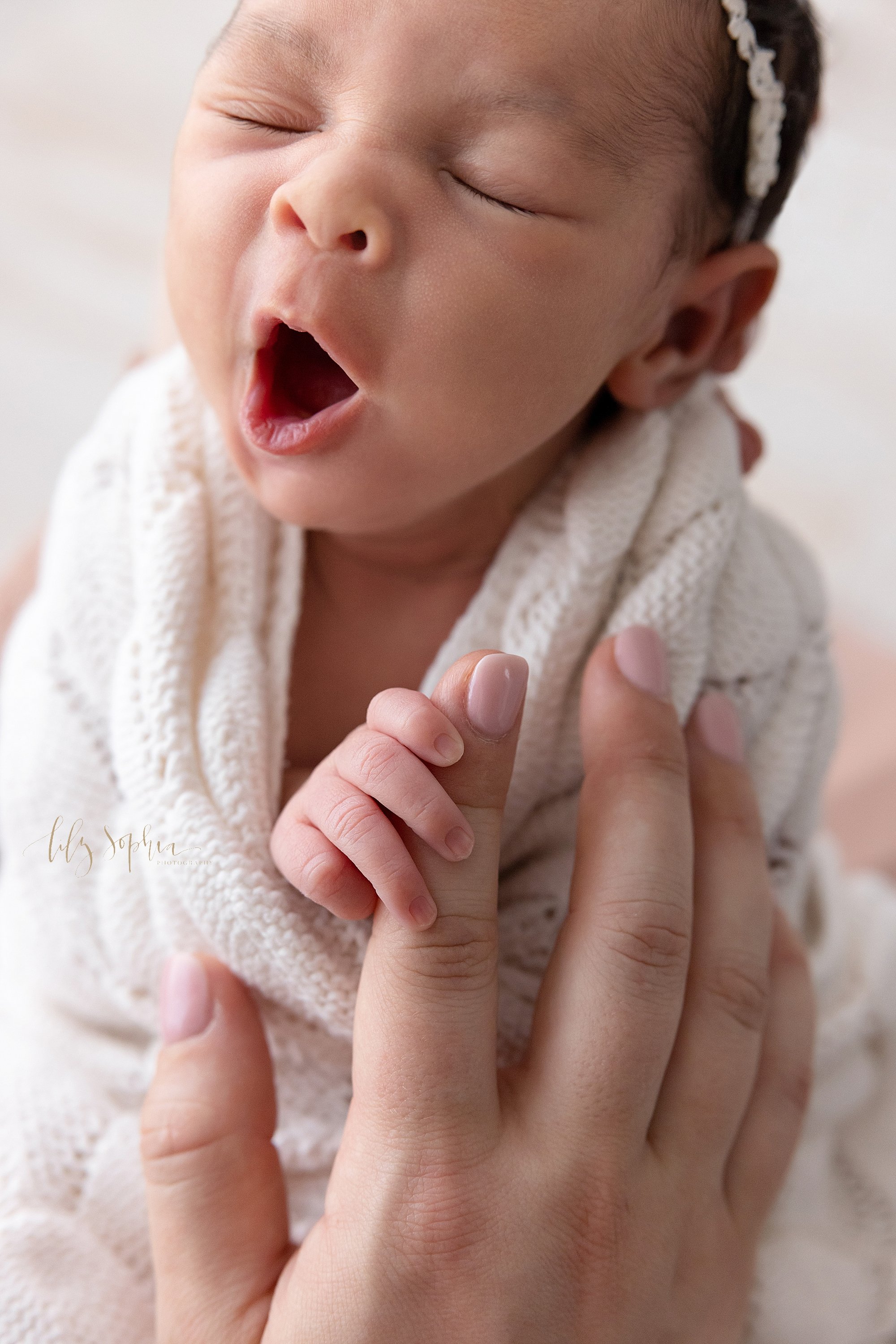  Close-up newborn portrait of a yawning newborn baby girl wearing a delicate headband in her black hair as she is wrapped in a soft white knitted blanket with her right hand peeking out to hold onto her mother’s index finger taken in a natural light 
