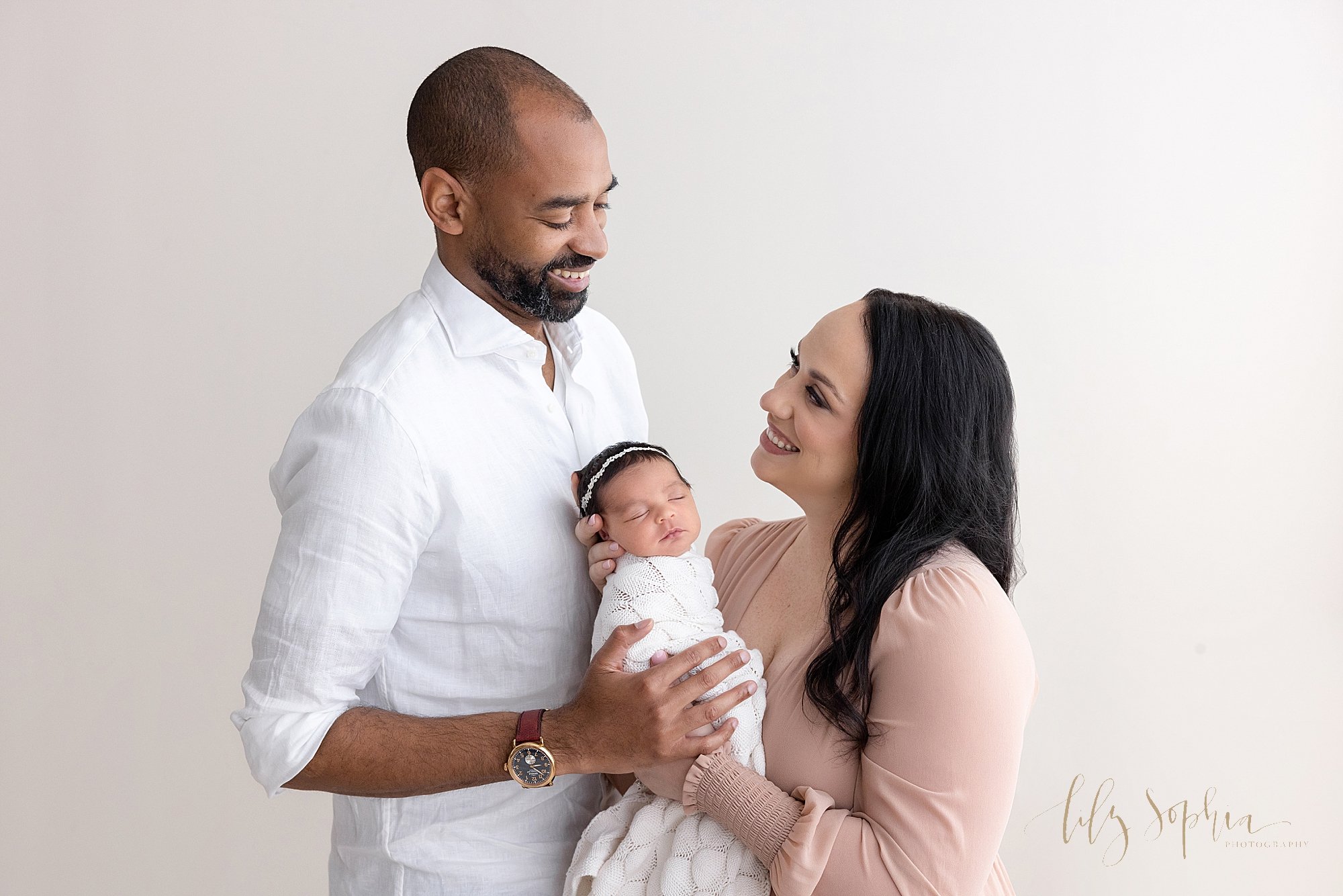  Newborn family photograph of a mother holding her newborn baby girl in her hands as she stands facing her husband and the two of them look lovingly at one another taken in natural light in a studio near Virginia Highlands in Atlanta. 