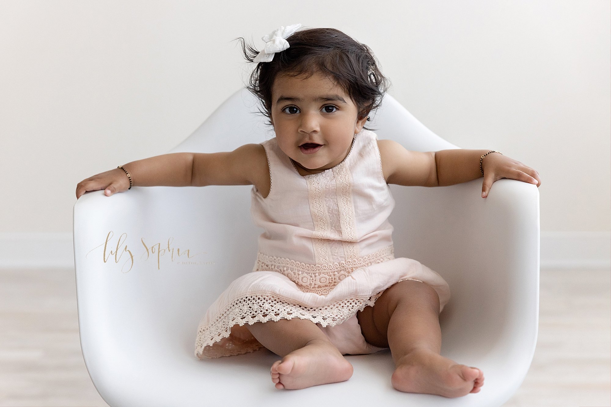  A proud one year old sits in a white molded chair to celebrate her first birthday in a studio using natural light as her picture is taken near Morningside in Atlanta, Georgia. 
